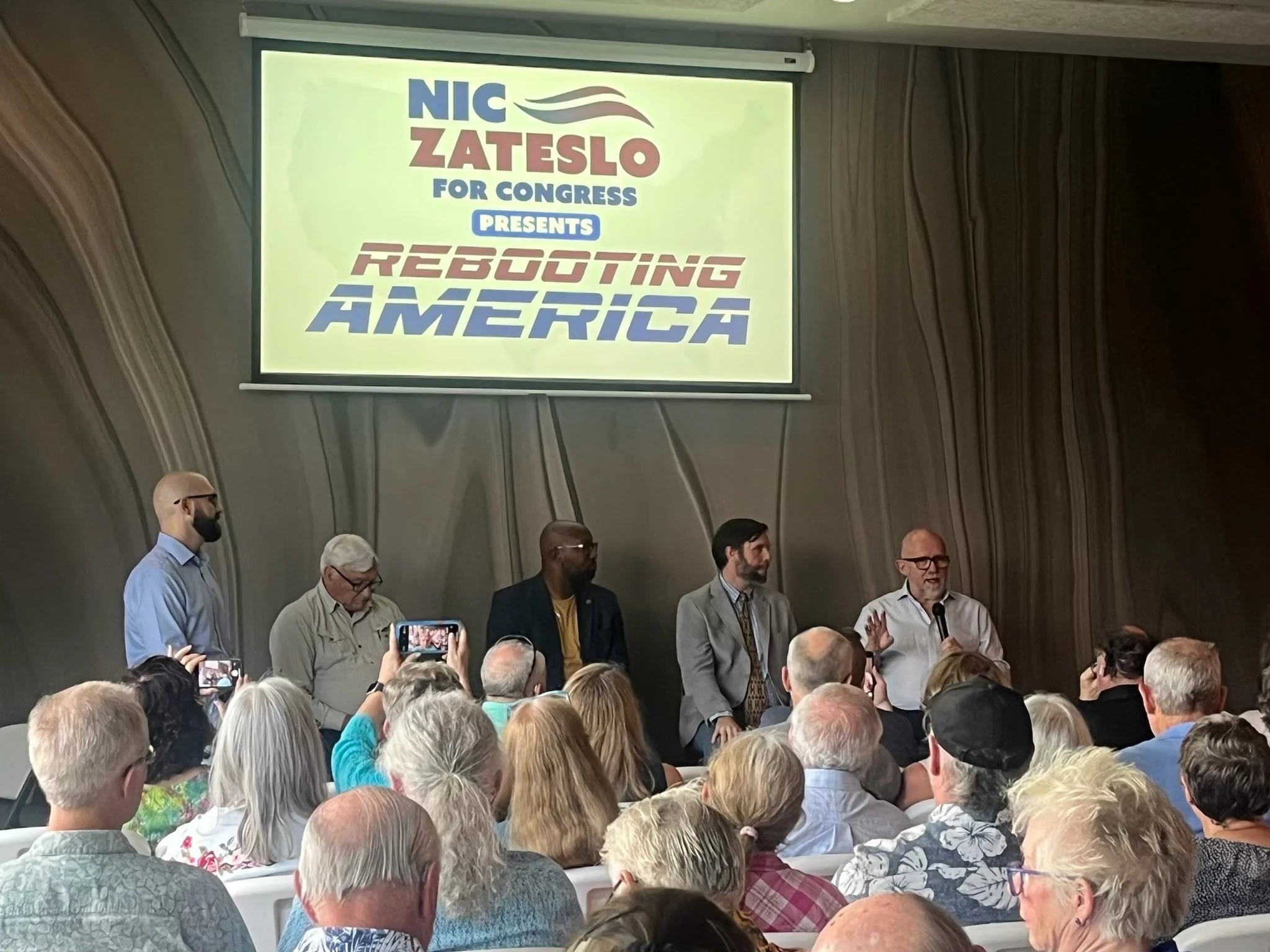 A panel discussion at an event with an audience, featuring a large screen displaying 'NIC ZATESLO FOR CONGRESS PRESENTS REBOOTING AMERICA' in the background.