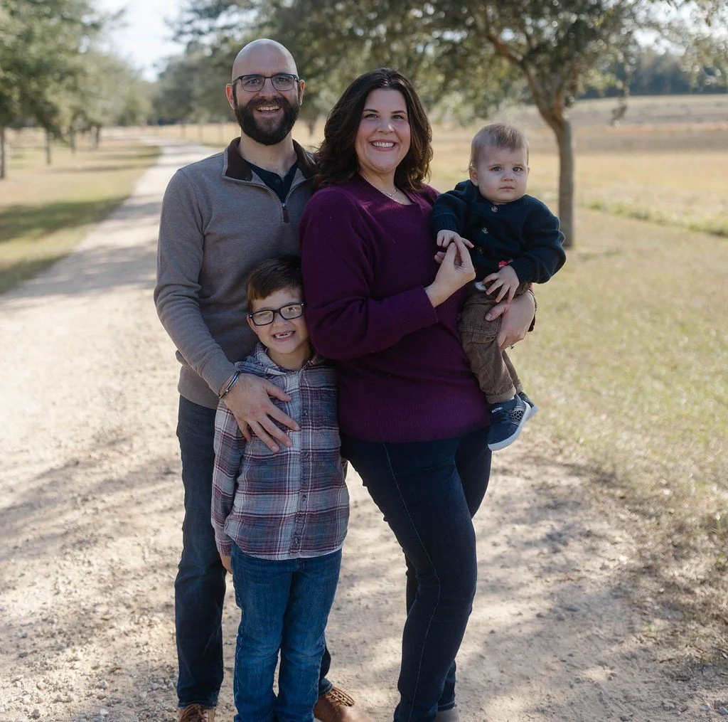 A family of four standing on a dirt pathway in a park on a sunny day with trees in the background. The mother is holding a toddler, and a boy with glasses is standing in front of them. The father stands behind the mother, smiling.