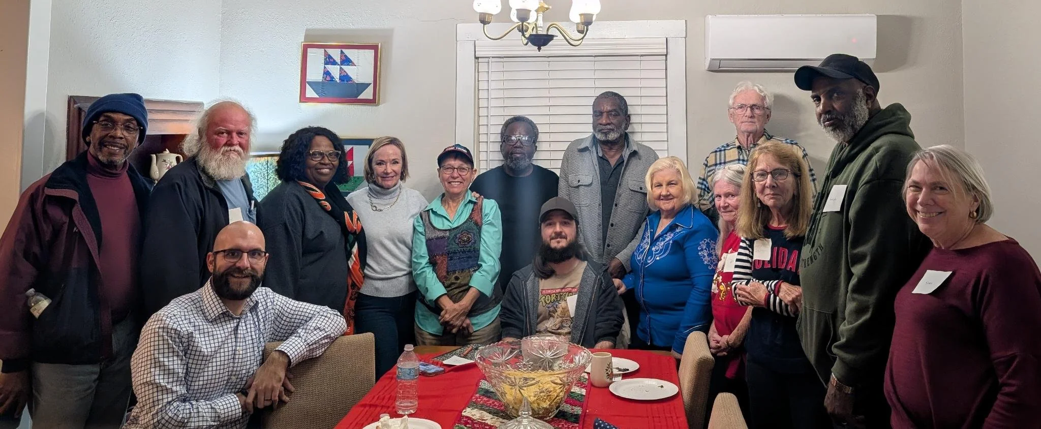 Group of diverse adults gathered around a table indoors, smiling for a group photo. The table has a large glass bowl, plates, and bottles, and the room features framed artwork and a window with closed blinds.