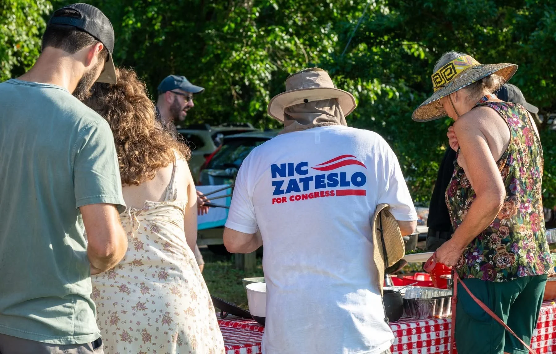 Group of people gathered outdoors at a table with a red and white checkered tablecloth, some wearing hats, around a person wearing a white shirt with the text 'NIC ZATESCO FOR CONGRESS'.