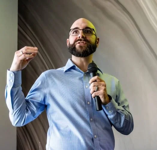 A man with a beard and glasses speaking into a microphone, wearing a blue dress shirt, gesturing with his left hand, standing in front of a wooden panel background.