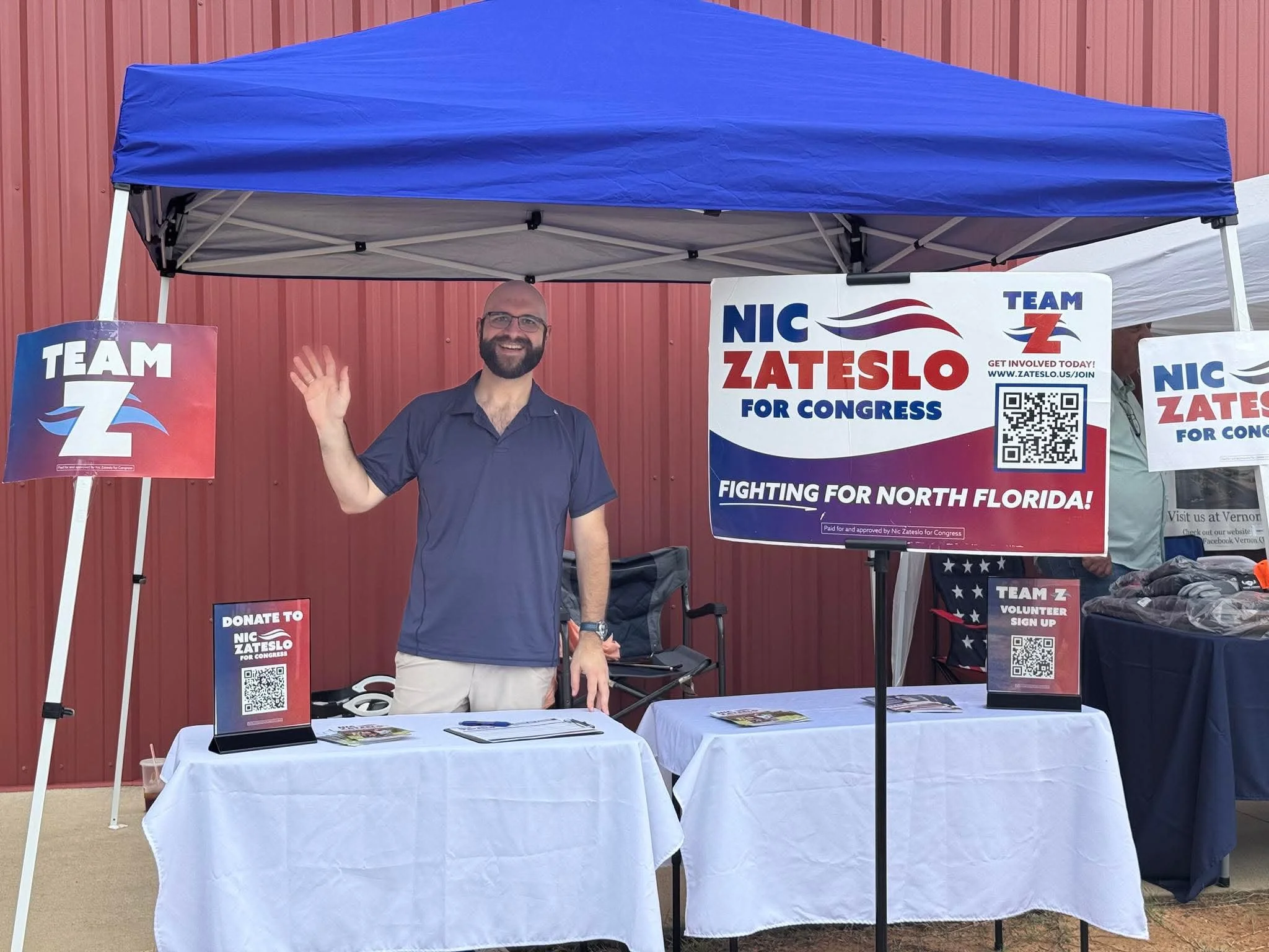 A man with glasses, a beard, and a navy blue shirt waving at an outdoor campaign booth for Nic Zateslo for Congress, with political signs and brochures on tables, set against a red wall.
