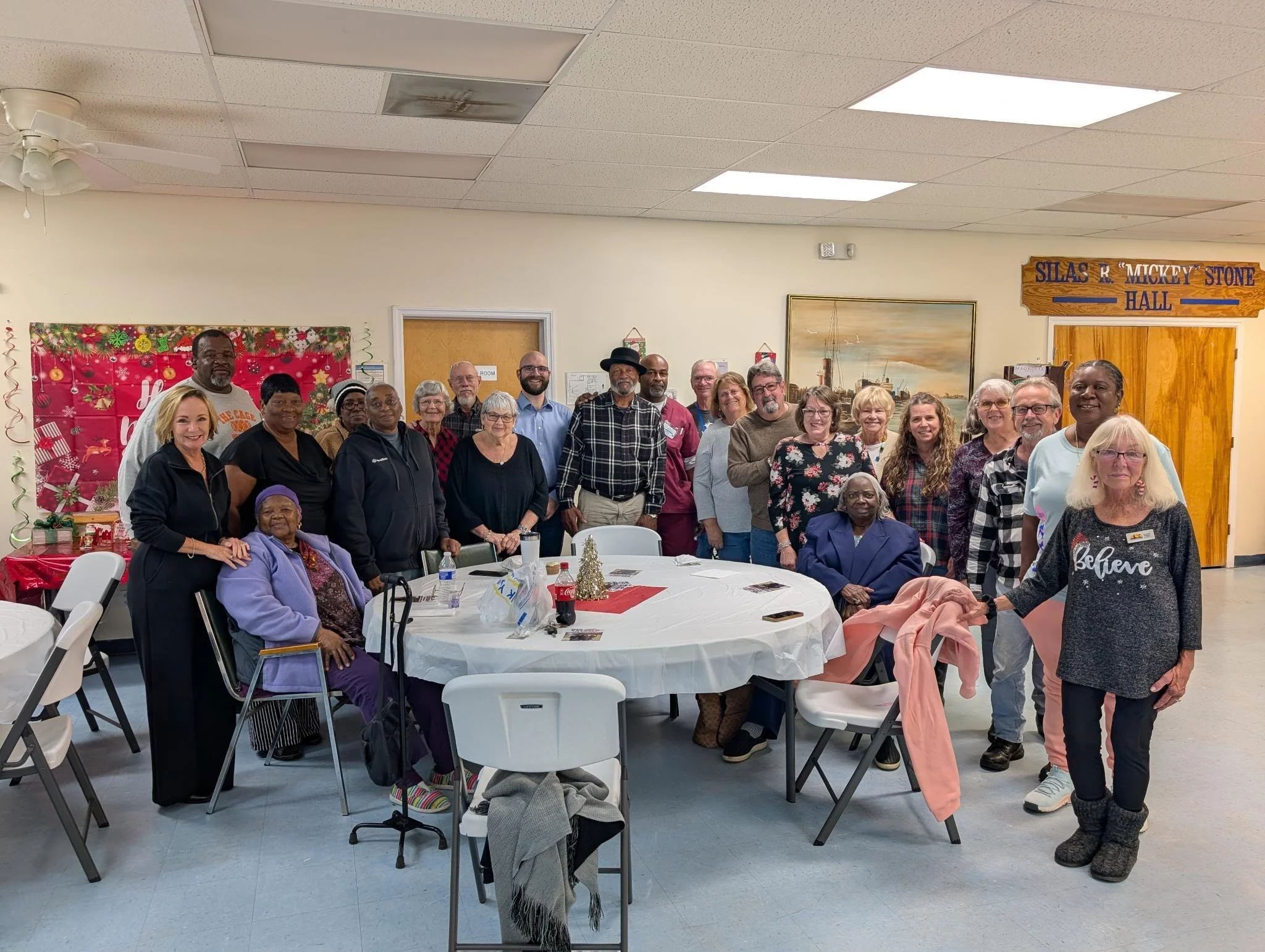 A group of people gathered around a decorated table in a community hall, celebrating a holiday event. There are about 20 adults, some standing and some seated, smiling at the camera. The room has holiday decorations, including a red and green backdro