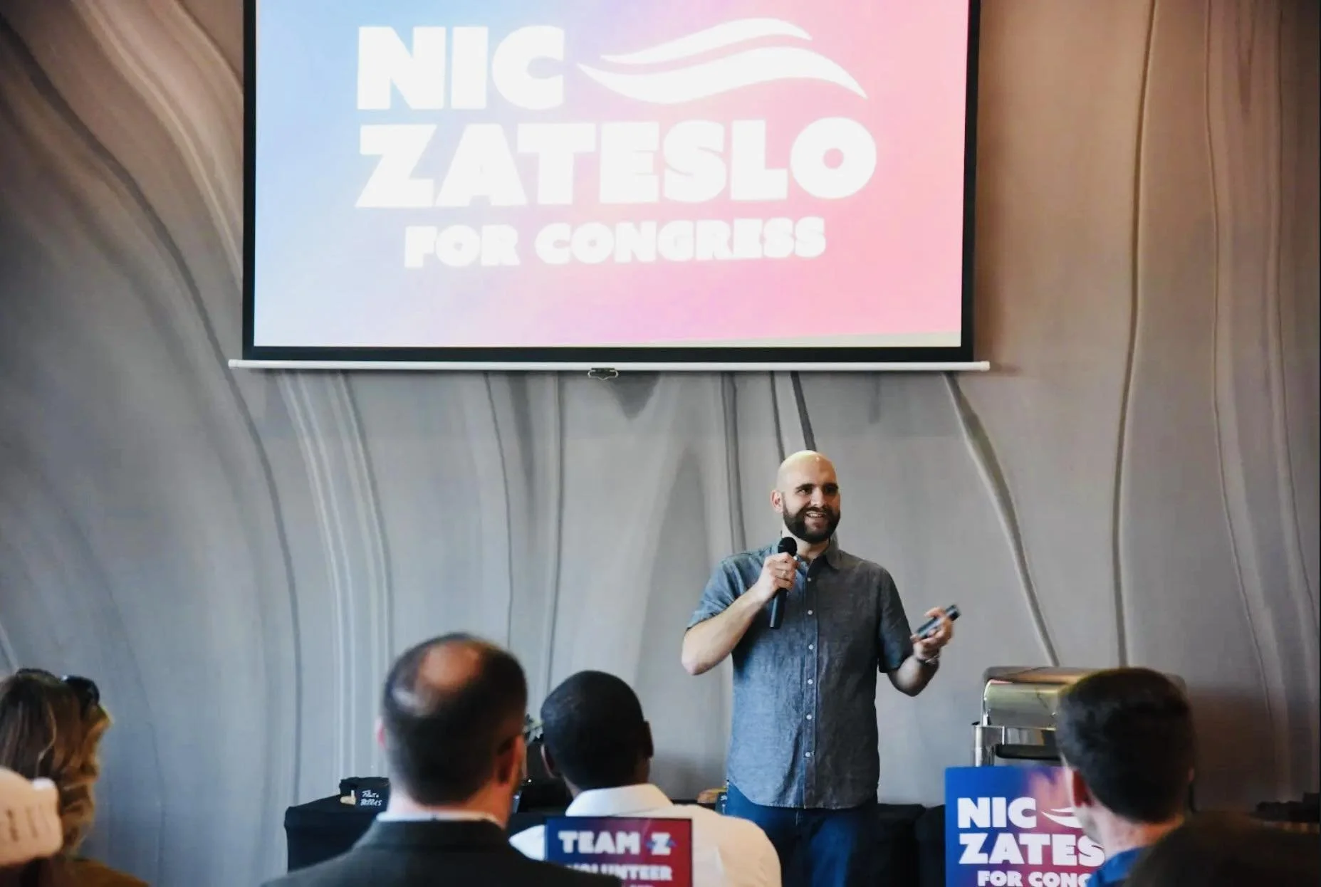 A man giving a speech in front of an audience at a political event with a large screen behind him displaying the name 'Nic Zateslo for Congress'.