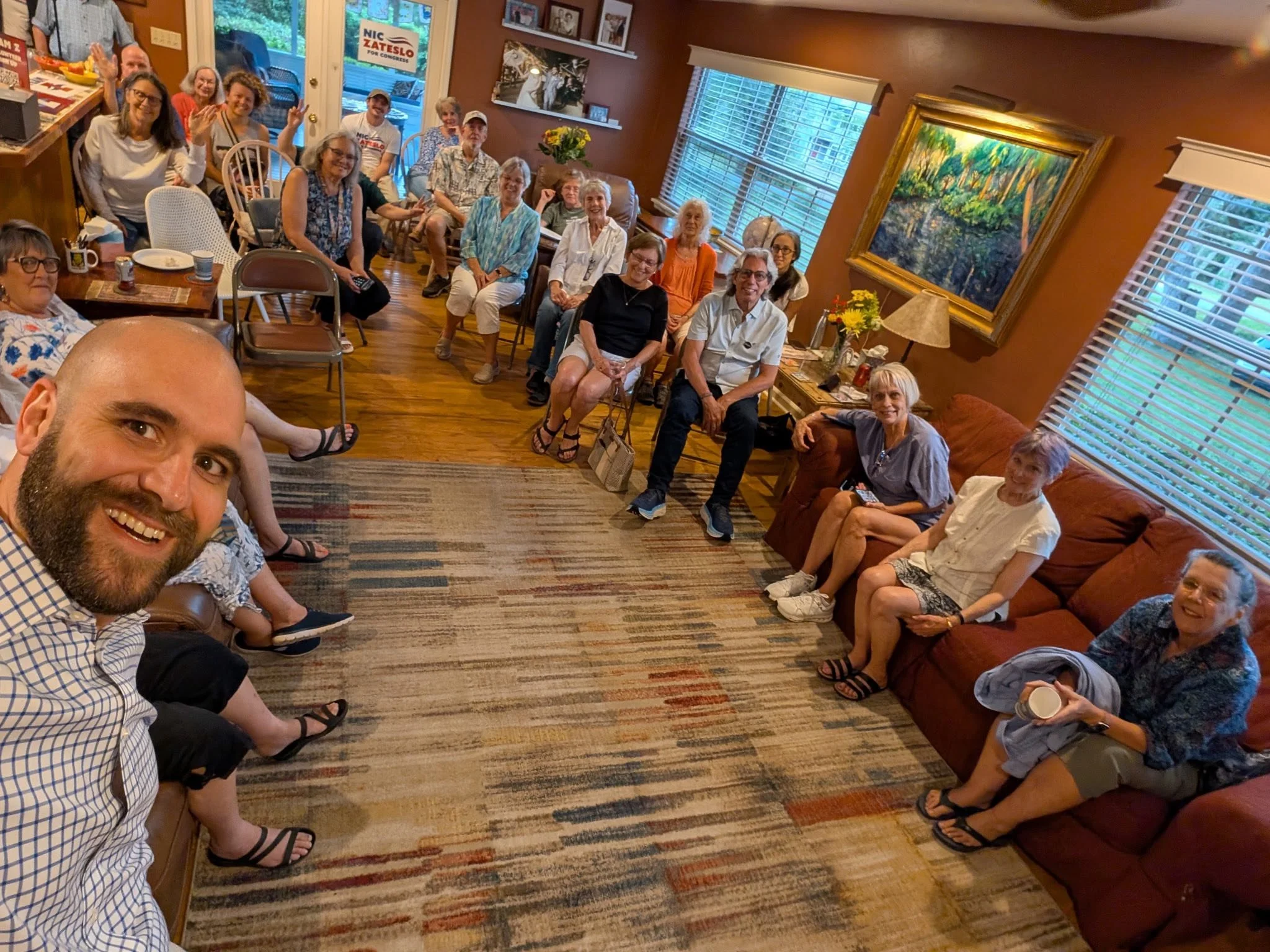 A group of people gathered in a cozy living room, sitting on chairs and couches, smiling and looking at the camera, with a man taking a selfie at the front left. The room has wooden floors, a patterned rug, large windows with blinds, and artwork on t