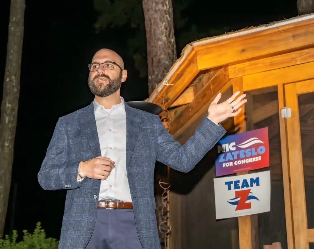 A man with glasses and a beard pointing at two political signs outside at night. One sign reads 'NIC Zateslo for Congress' and the other 'Team Z'. The signs are attached to a wooden structure, and the background includes trees and darkness.
