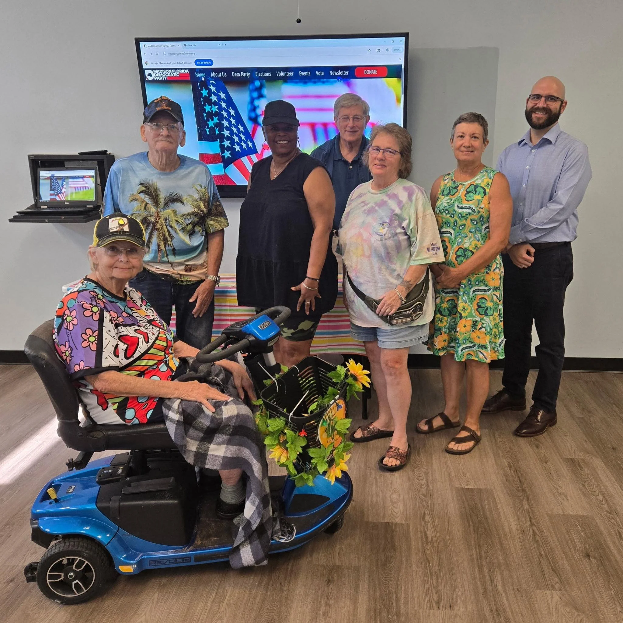 A group of seven people posing for a photo indoors, with a woman in a motorized scooter decorated with flowers in the foreground. The woman is elderly and wearing a colorful shirt and plaid blanket. Behind her, there are six adults standing in front 