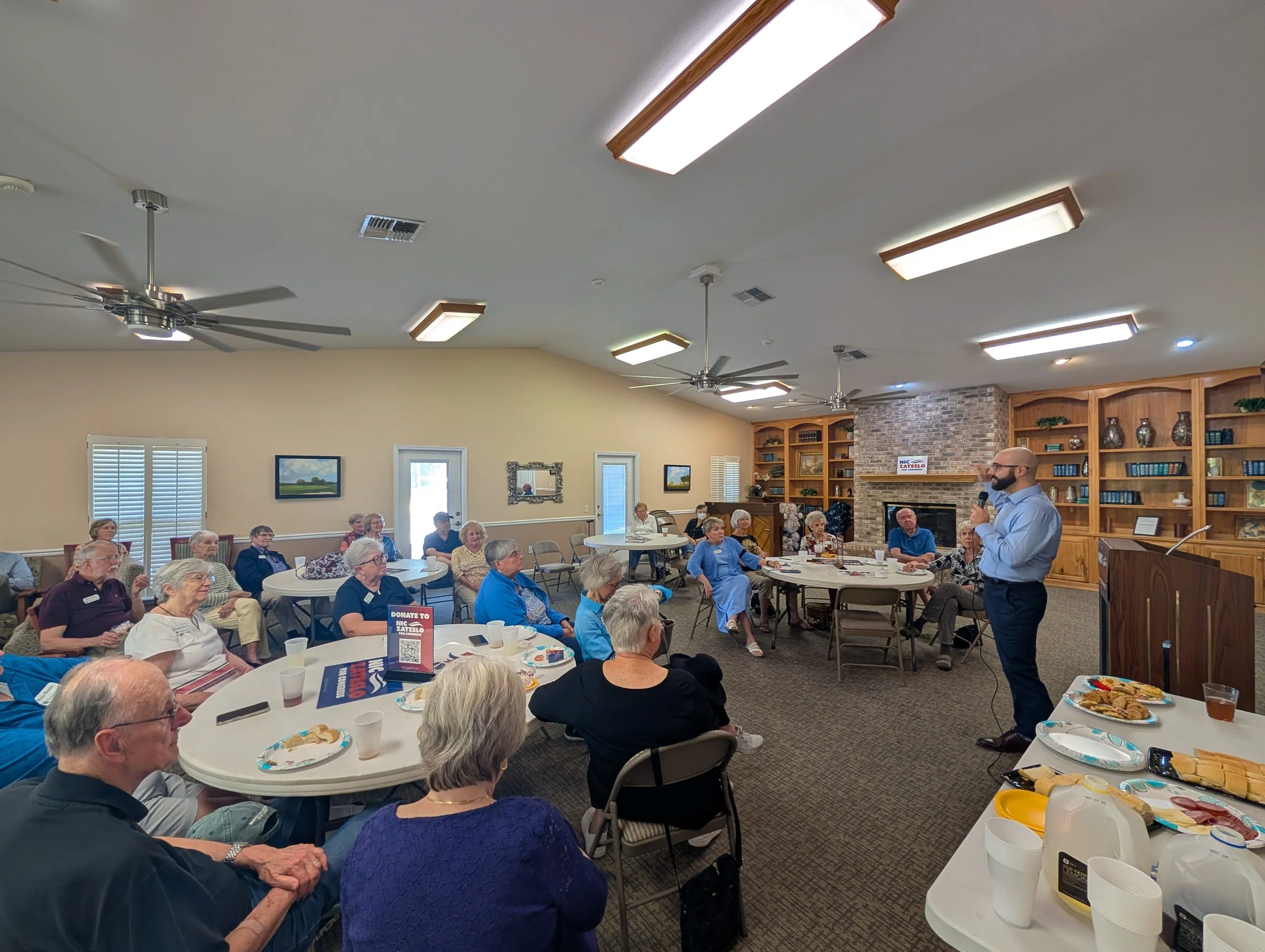 A man giving a presentation to a group of elderly people in a community room with tables, snacks, and bookshelves.