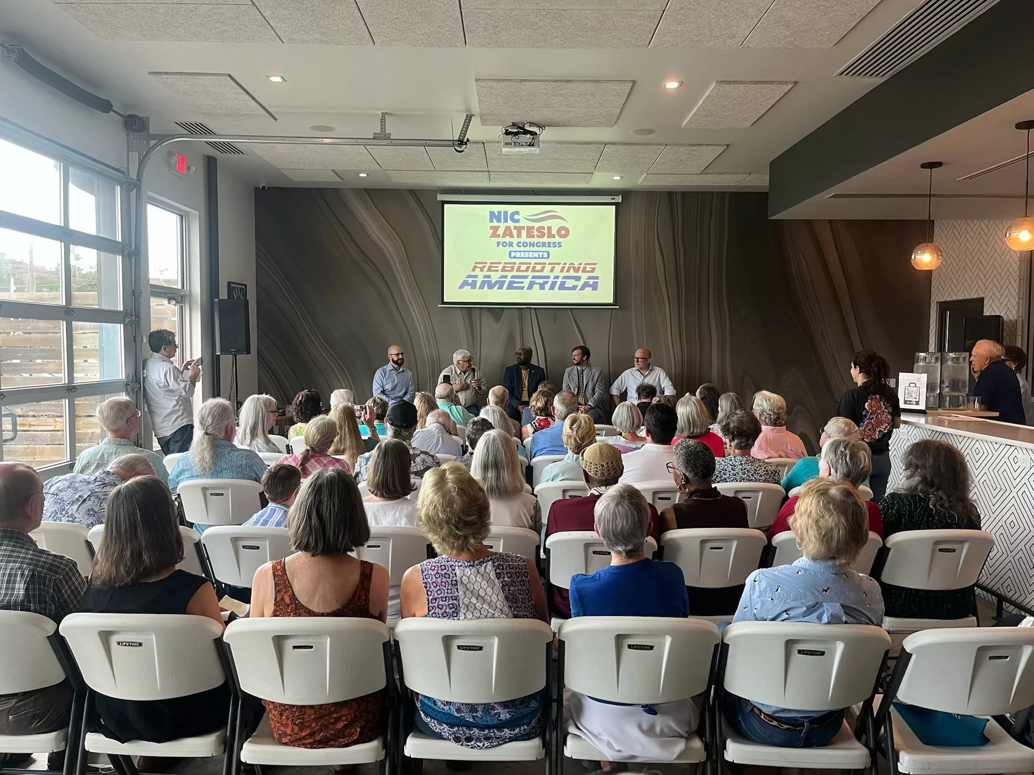 A large indoor conference room filled with a diverse audience watching a panel of six speakers on stage. The screen behind the speakers displays a political campaign message, with the text 'NIC ZATESLO FOR CONGRESS PRESENTS REBOOTING AMERICA.' The ro
