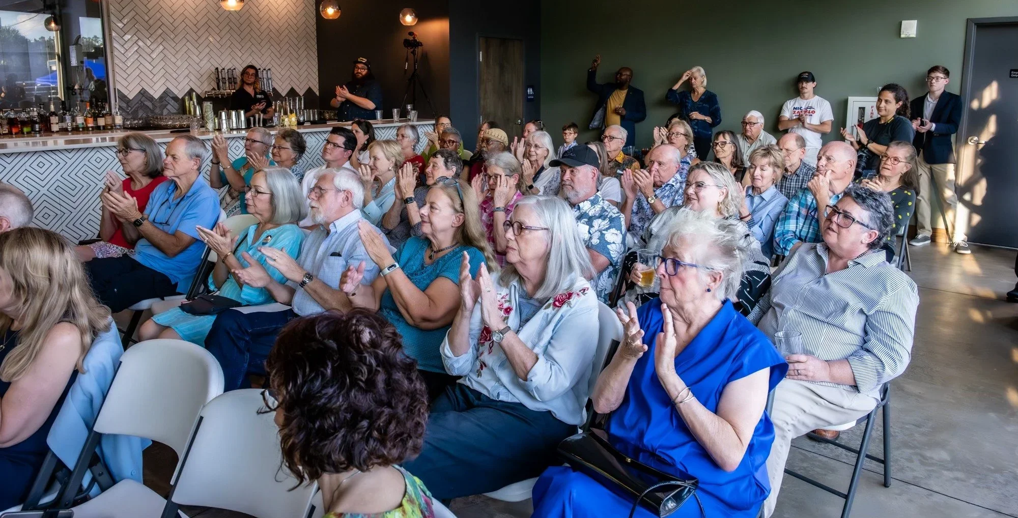 A large group of mostly elderly people are sitting and standing in a restaurant or event space, attentively watching or listening to something, with some clapping and others raising their hands. The room has modern decor, a bar area on the left, and 