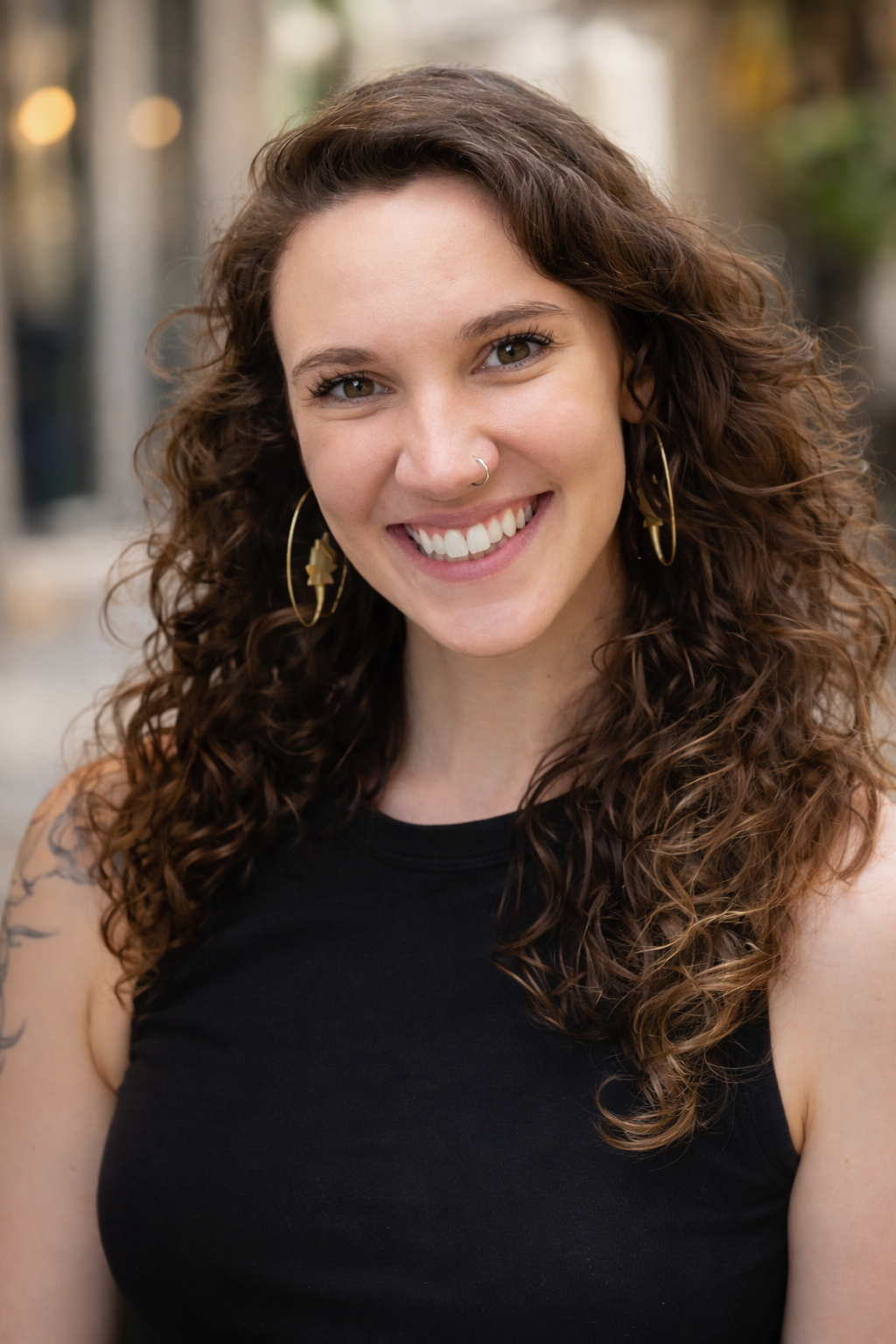 A young woman with long, curly brown hair smiling outdoors, wearing hoop earrings and a black sleeveless top.