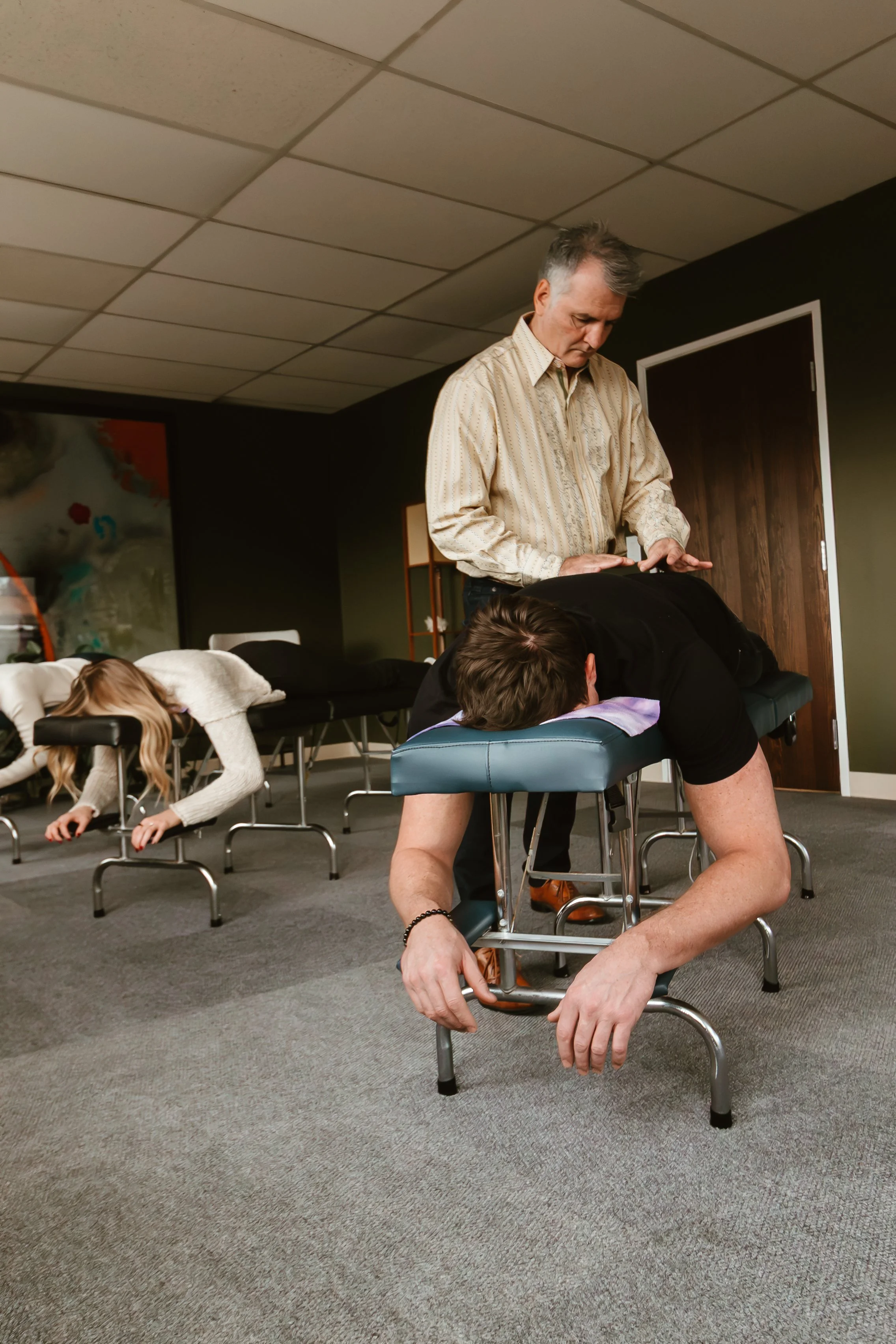 A person lying face down on a massage table receiving a back massage from a therapist, with a young girl lying face down on an adjacent table in a room with dark walls and a colorful painting.