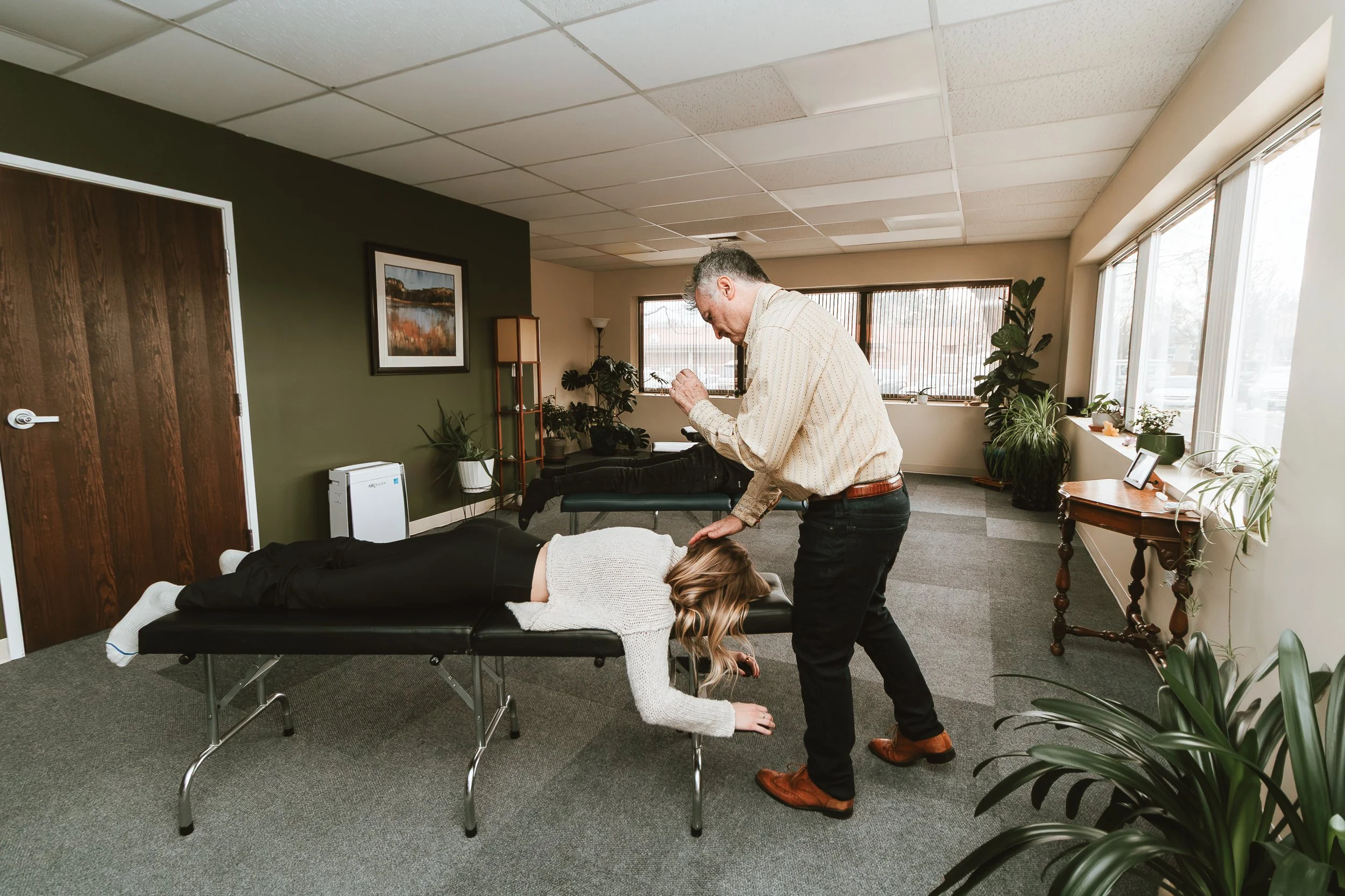 A chiropractor treats a woman lying face down on a massage table in an office, with another patient in the background and various potted plants around.