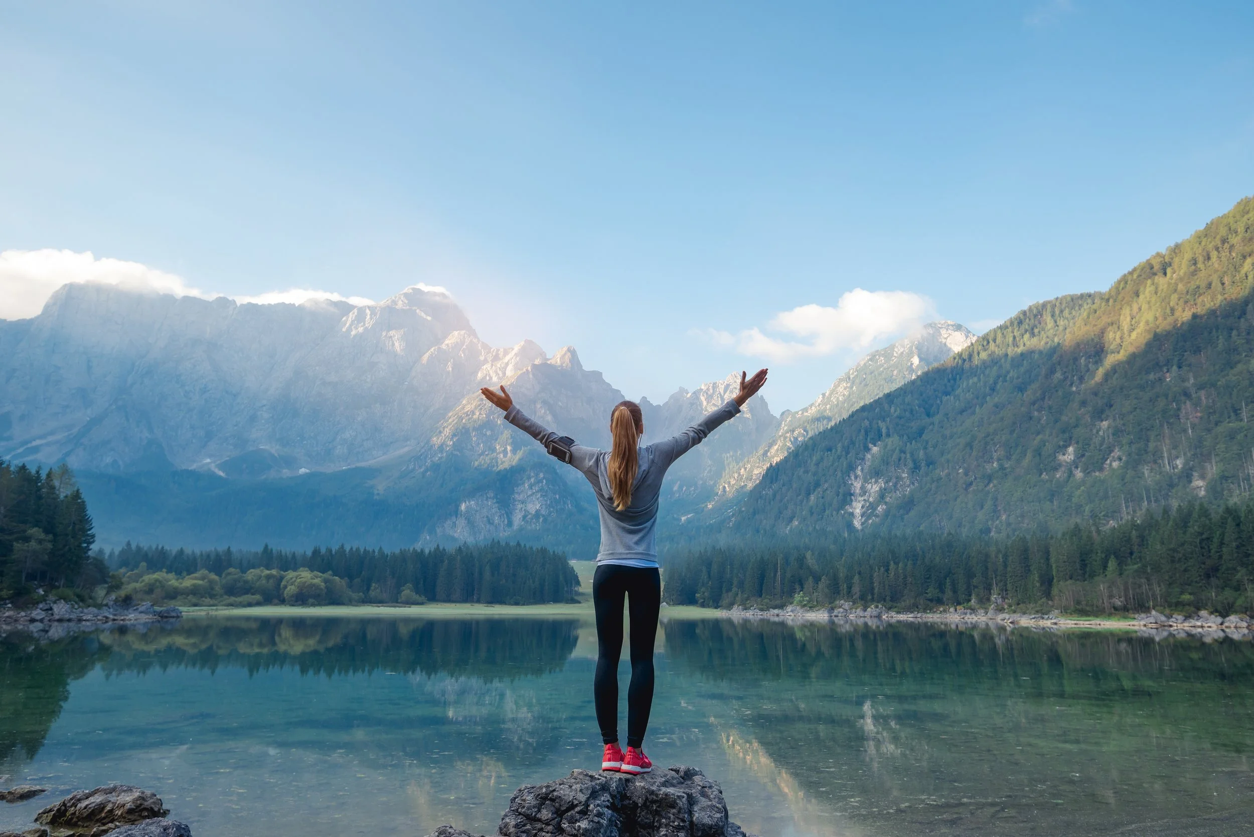 Woman standing on a rock with arms raised, looking at a mountain lake surrounded by forested mountains under a blue sky.