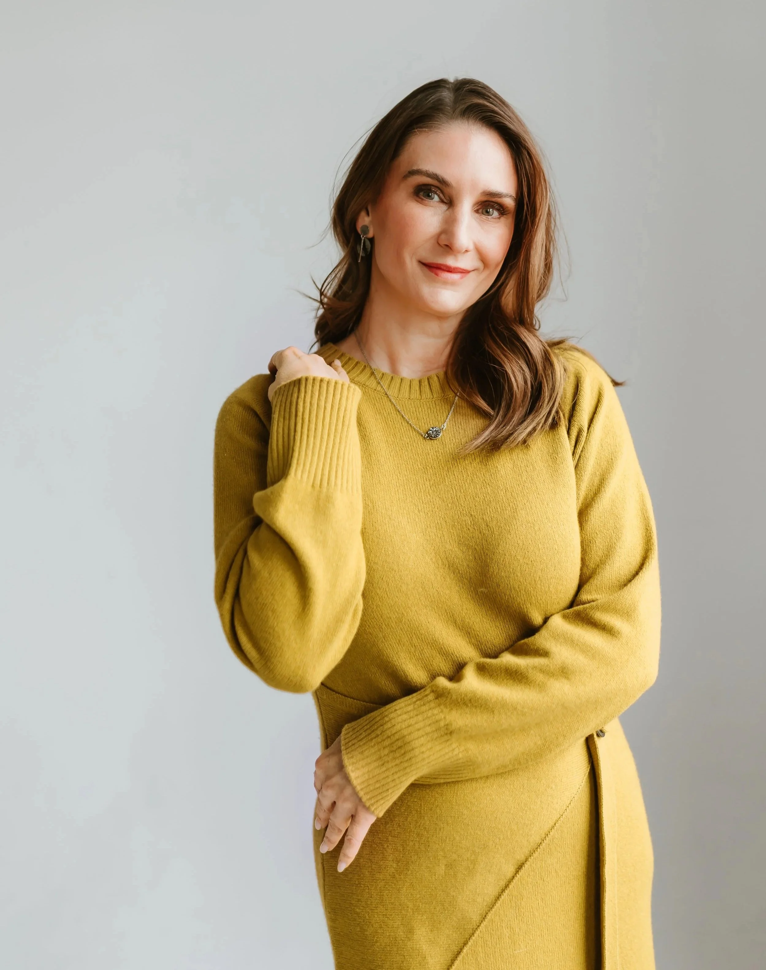 A woman wearing a yellow sweater and skirt, standing against a plain light gray background, with brown hair and accessories including earrings and a necklace.