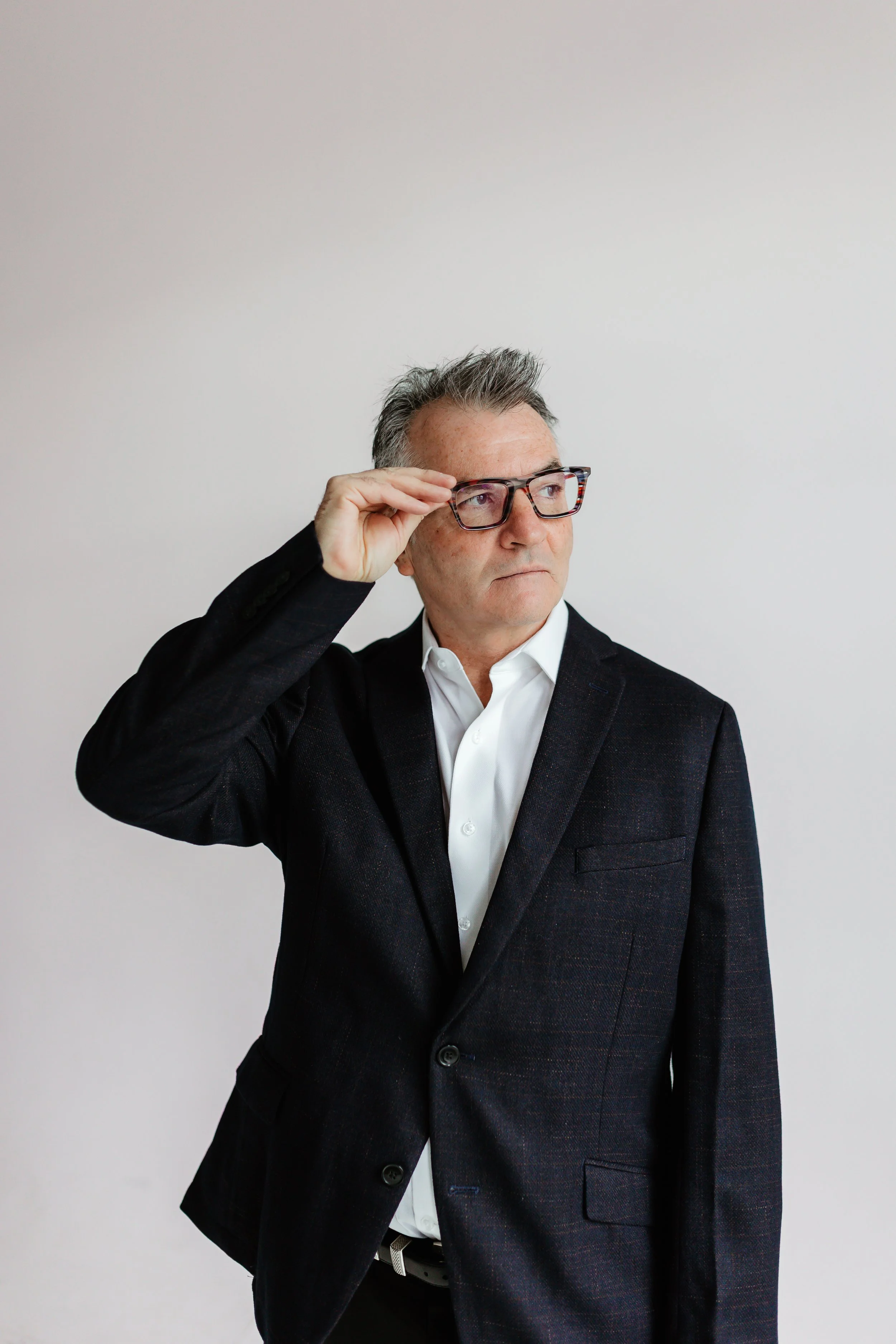 An older man in a black blazer and white shirt, wearing glasses, is looking to the side and slightly lifting his glasses with his right hand, against a plain light background.
