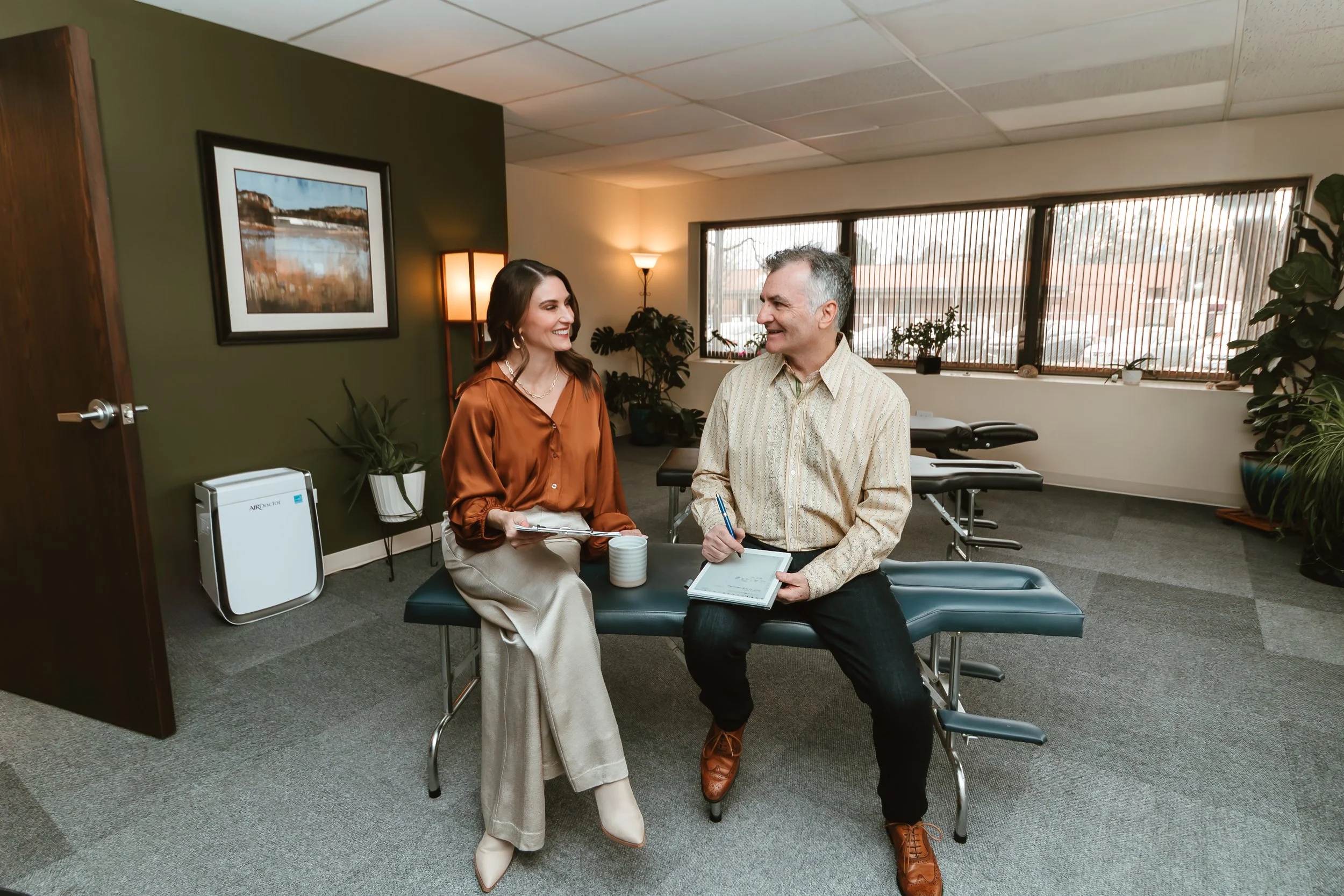 A woman and a man sitting on a therapy table in a therapy or chiropractic clinic, smiling and talking. The woman holds a clipboard, and the man has a notepad and pen. The room has large windows with blinds, plants, and framed artwork on the wall.