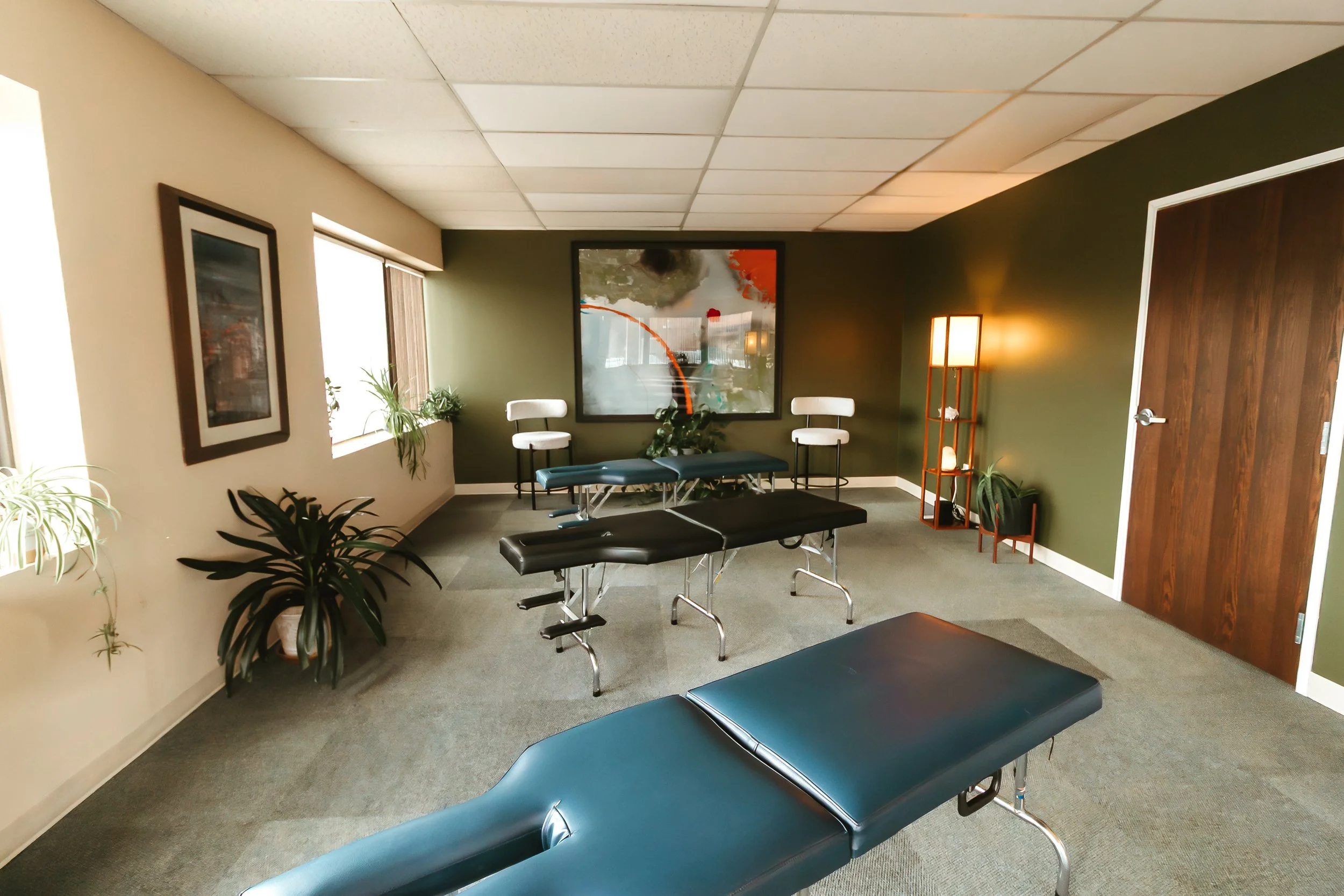 Massage therapy room with two massage tables, one black and one teal, in a space with green and beige walls, artwork, potted plants, and a wooden door.