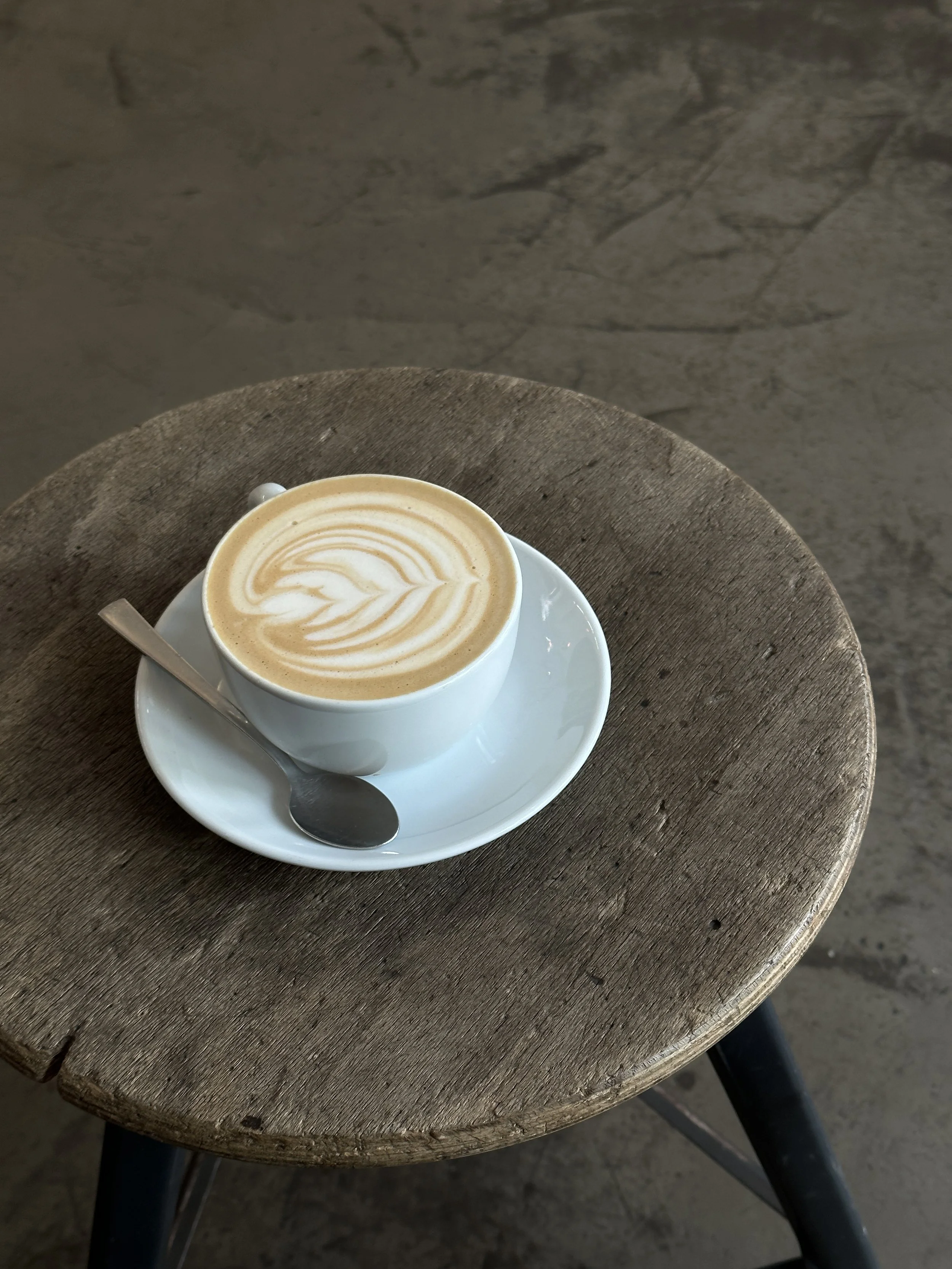 A white cup of latte with latte art on top, placed on a matching white saucer with a metal spoon, on top of a rustic wooden round table.