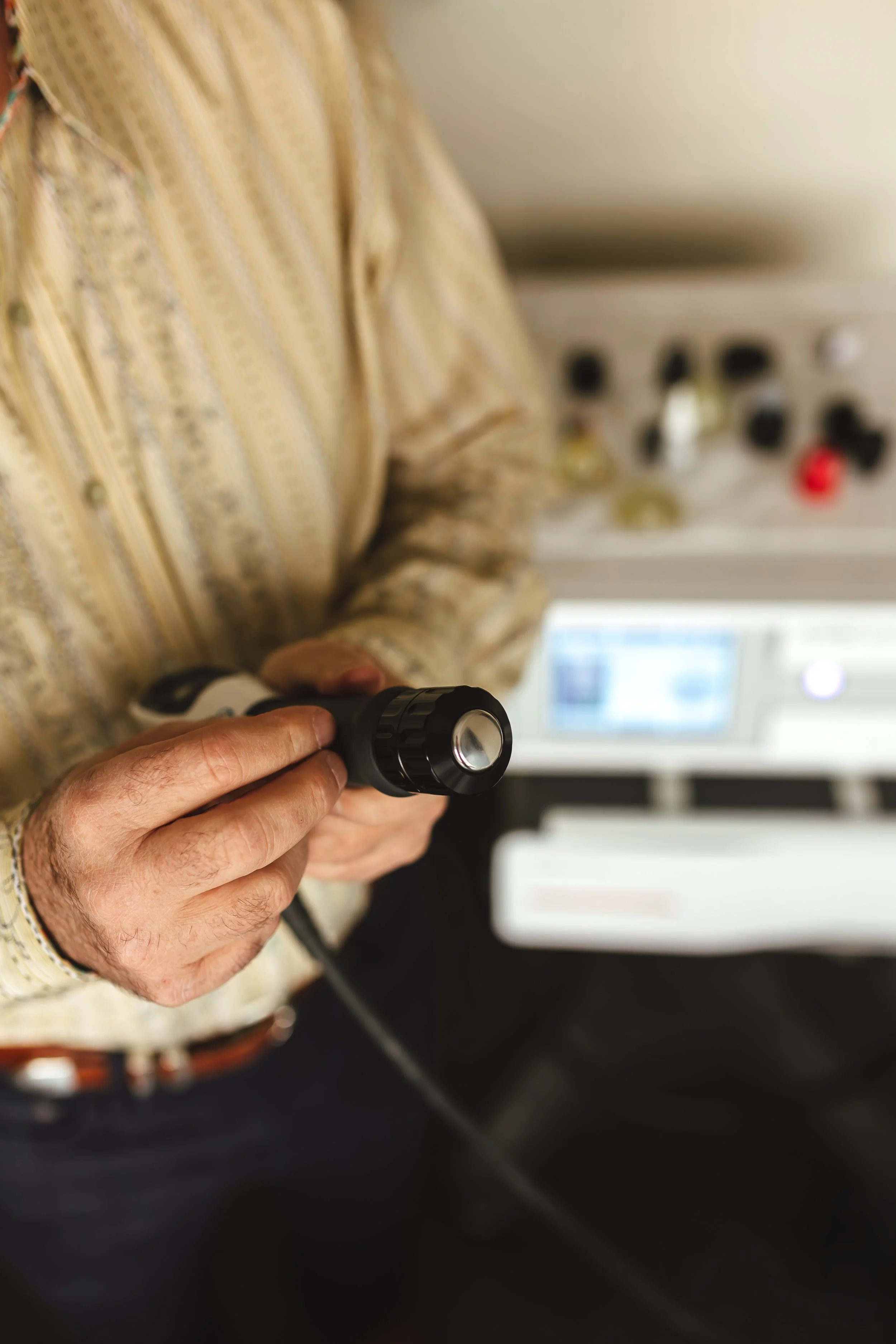 Close-up of an older person's hands holding a black flashlight, with a computer and out-of-focus electronic devices in the background.