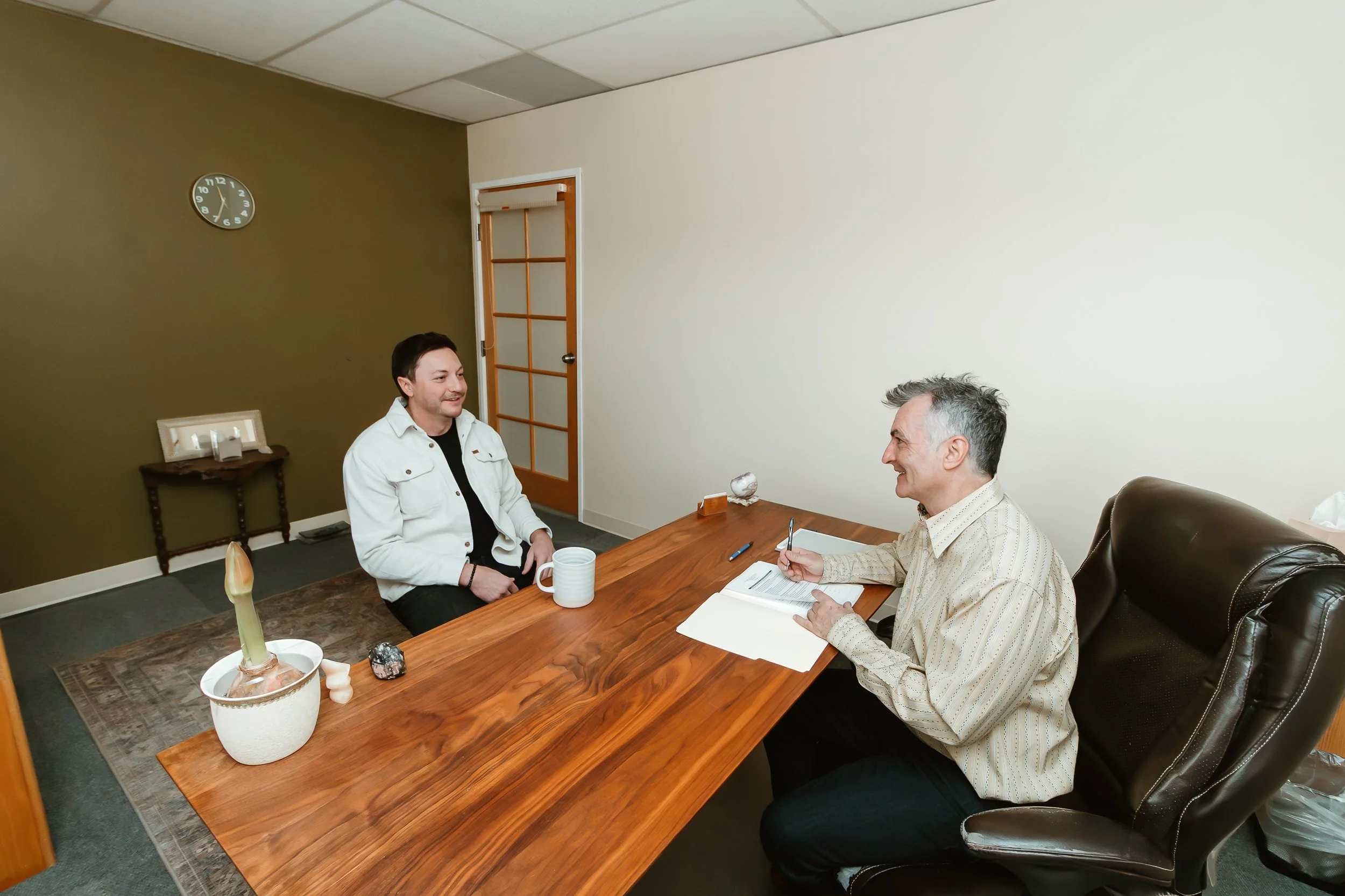 A man and a woman sitting across a wooden table in an office or consultation room. The woman, on the left, is wearing a white jacket and is smiling. The man, on the right, is wearing a light-colored striped shirt, holding a pen, and smiling. There are notebooks, a mug, and decorative objects on the table. The room has a clock on the wall and a door with glass panes.