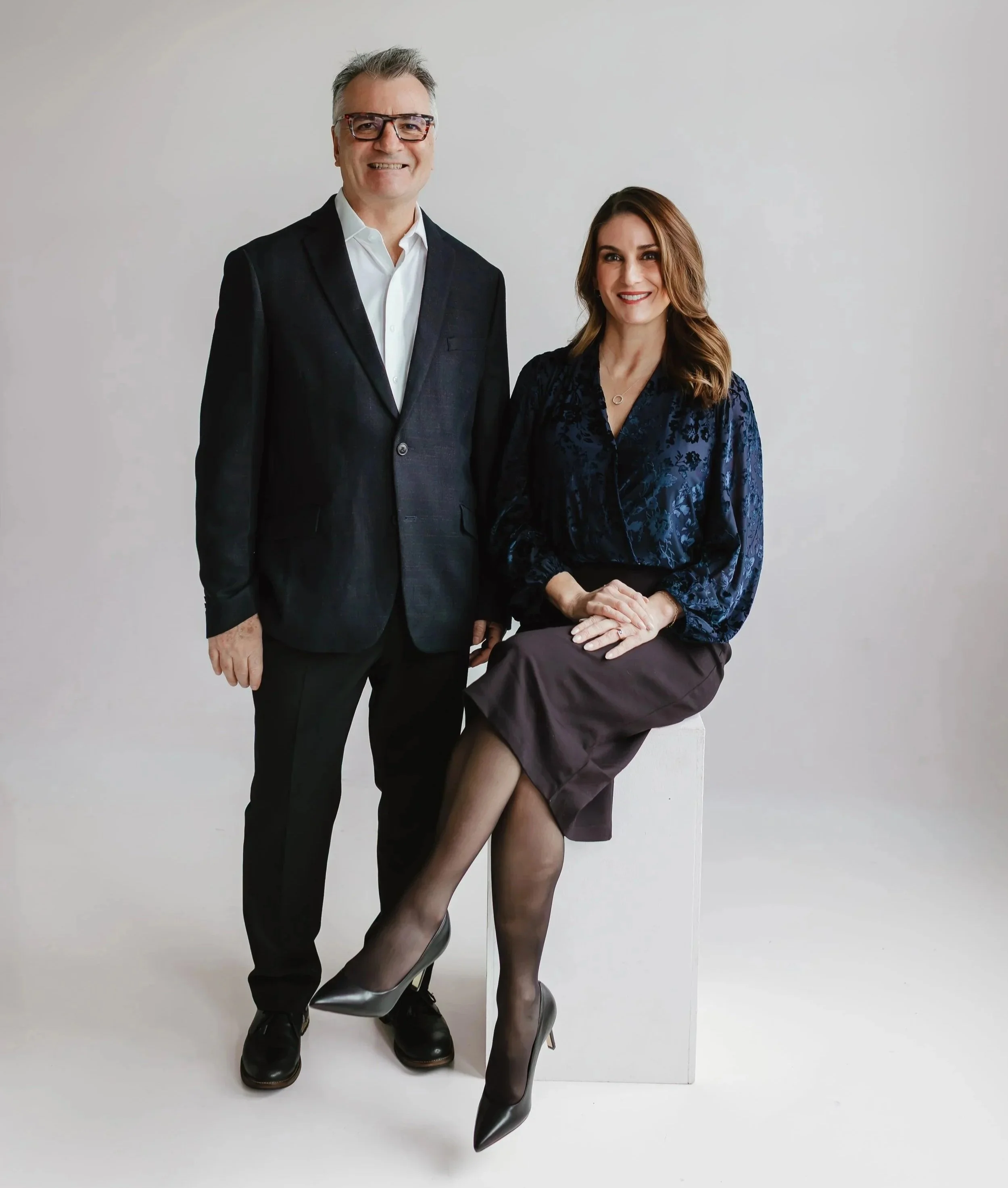 A man in a dark suit and glasses standing next to a woman in a navy blouse and brown skirt sitting on a white block, against a plain light-colored background.