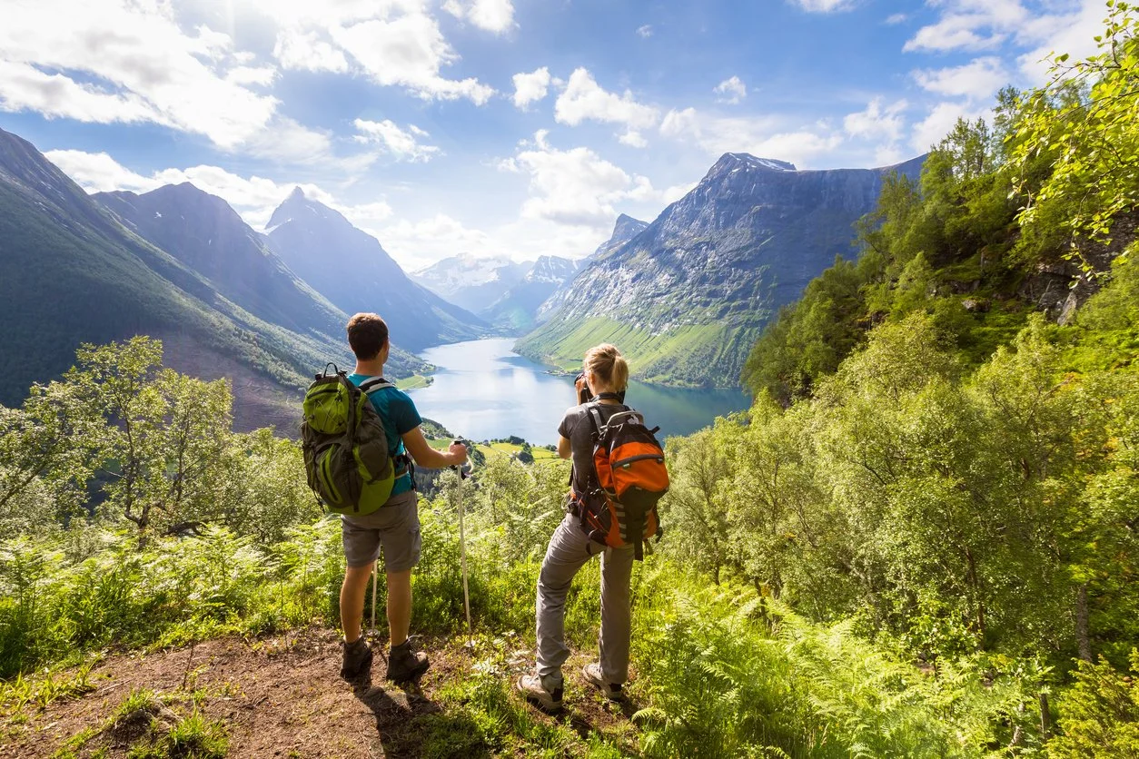 Two hikers overlook a scenic lake in a mountainous valley surrounded by green trees and snow-capped peaks under a partly cloudy sky.