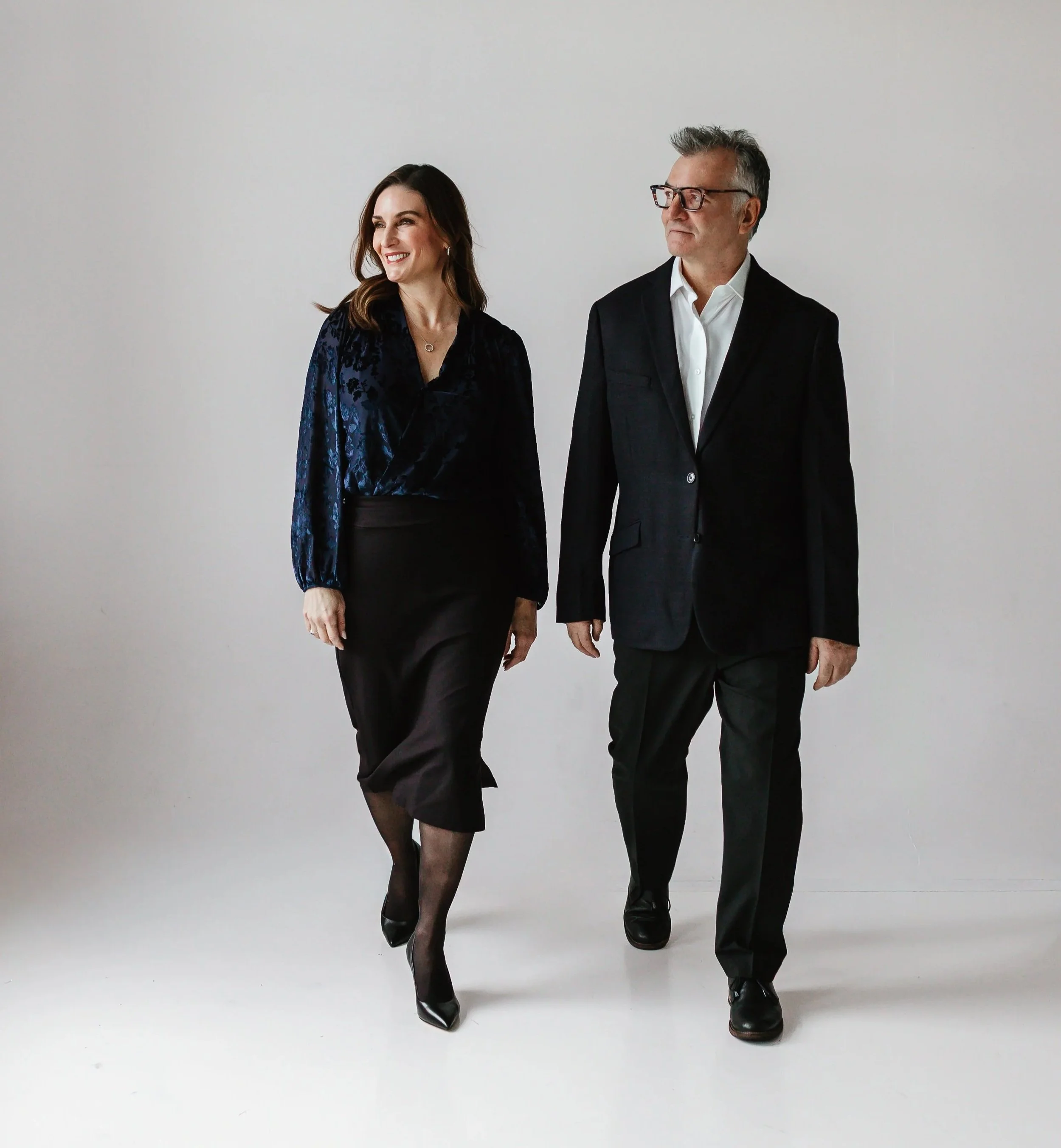 A woman and a man, both dressed in formal black attire, walking together against a plain white background.
