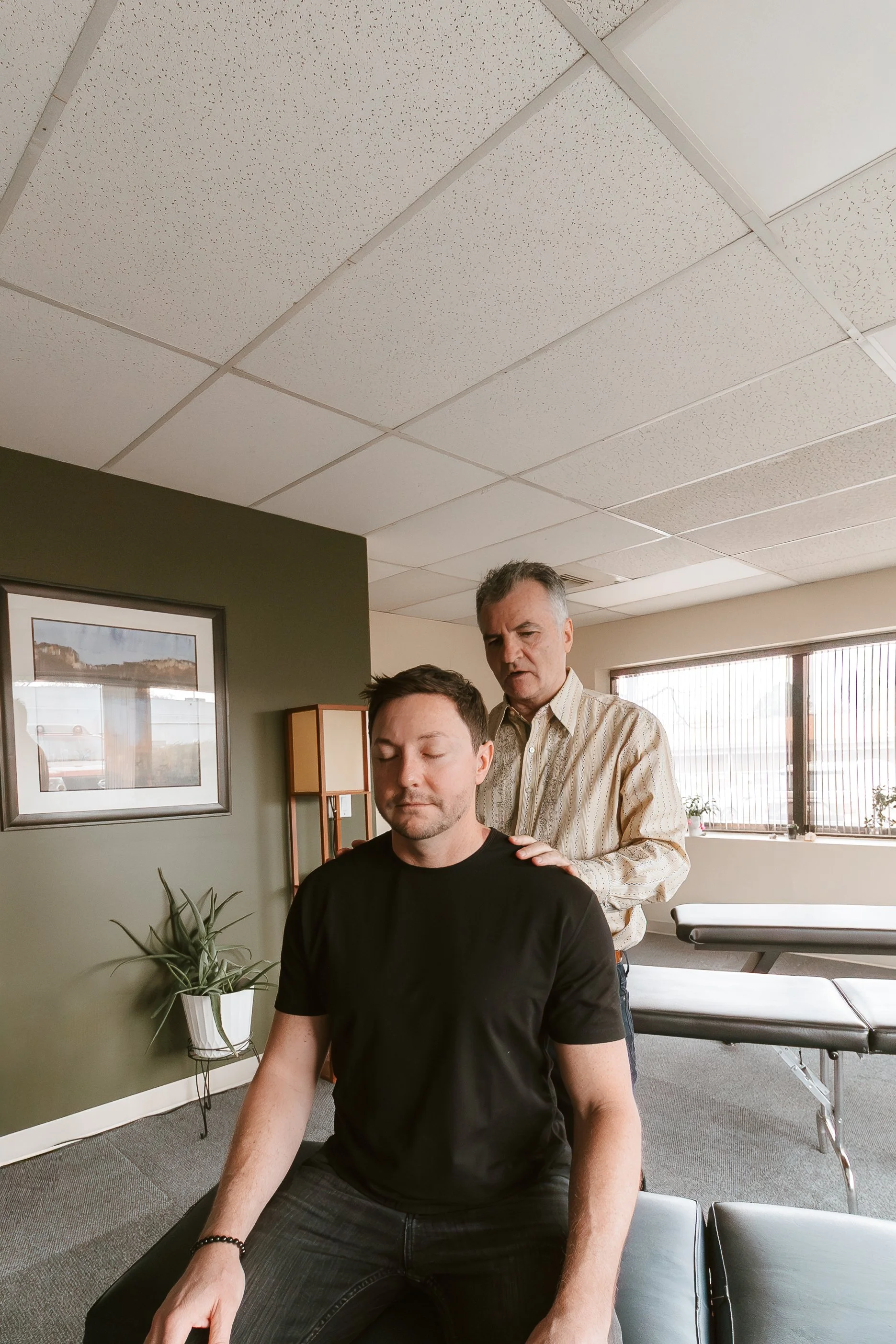 A man in a black T-shirt sitting with eyes closed, receiving a massage from a man standing behind him in an office or therapy room with a green wall, a framed picture, a potted plant, and large windows.