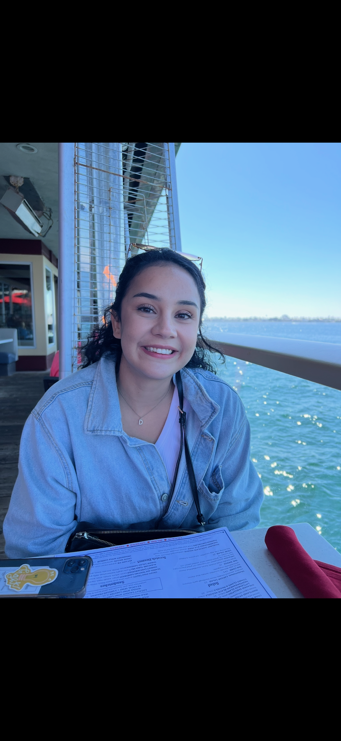 A young woman with dark hair wearing a light denim jacket, smiling while sitting at a table near the water with a menu, a red rolled napkin, and a smartphone with a sticker of a yellow cartoon character.