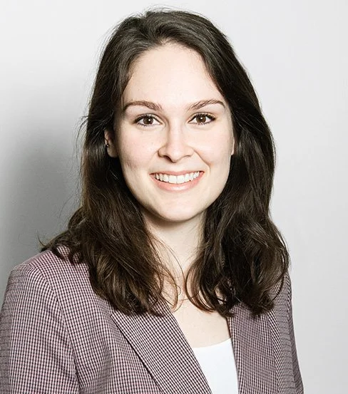 Portrait of a young woman with long brown hair, smiling, wearing a checkered blazer and a white top, against a plain light gray background.