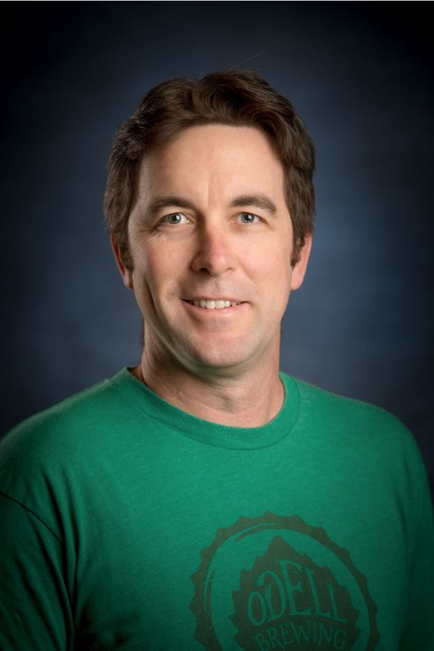 A man with brown hair wearing a green t-shirt smiling at the camera, against a dark background.