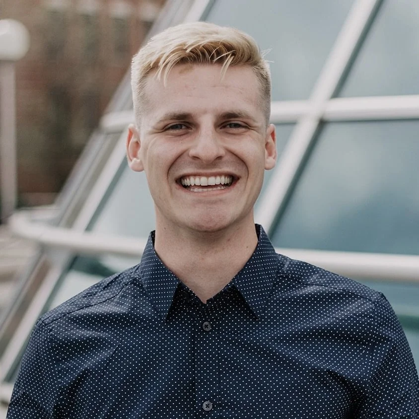 Young man smiling outdoors with modern glass building in background.