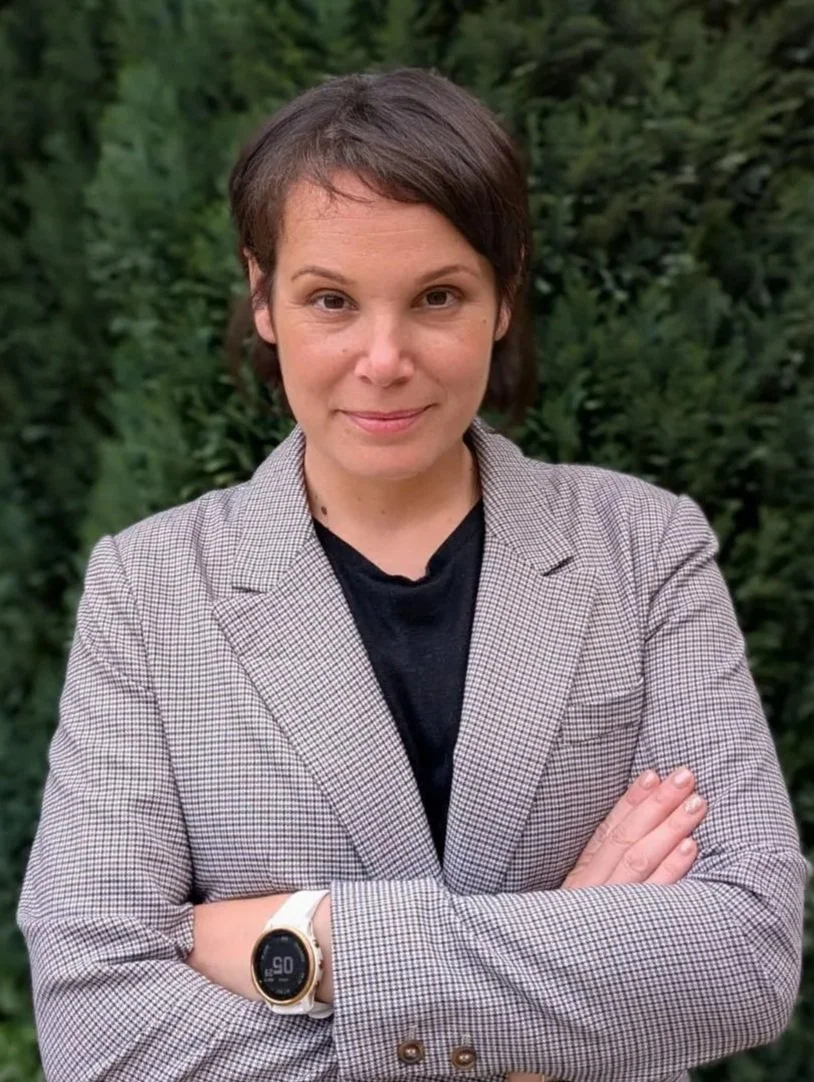 A woman with short brown hair standing outdoors with crossed arms, wearing a checked blazer and a black top, in front of green foliage.
