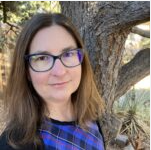 A woman with long brown hair, glasses, and a blue plaid shirt standing outdoors next to a large tree.