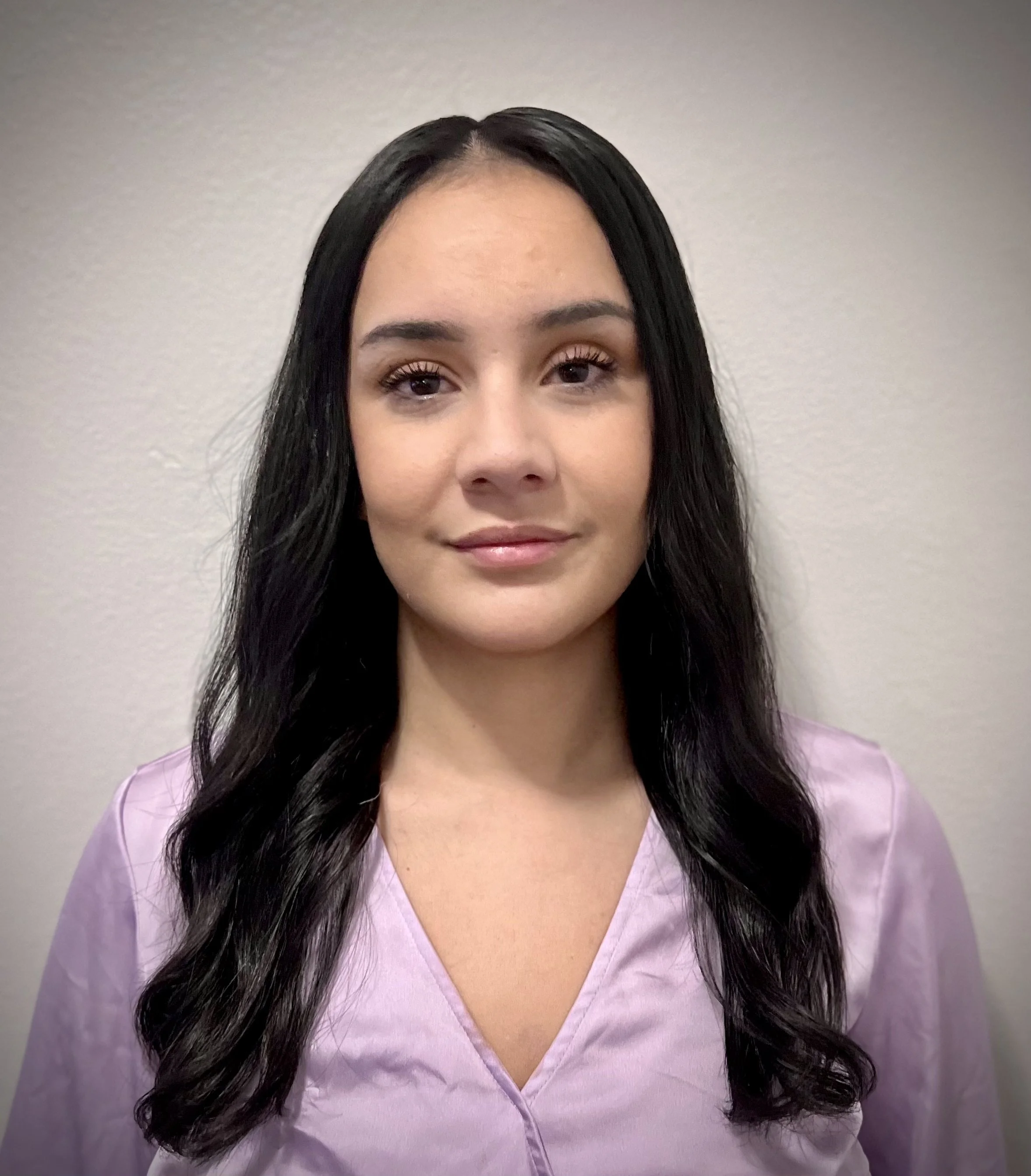 A young woman with long black hair wearing a light purple medical scrub top, standing against a plain beige wall.