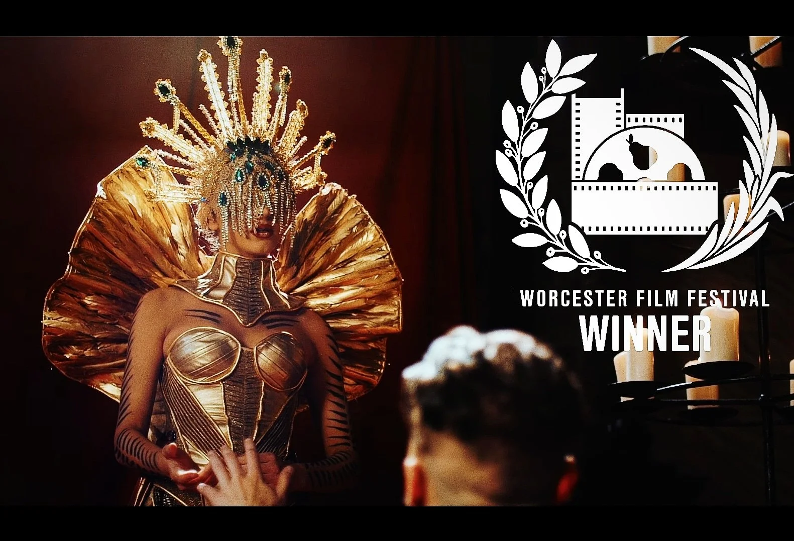 A performer in an elaborate gold costume with a large fan-like collar, gold headdress with jewels, gold face mask, and black striped body paint, standing in front of a dark background with a logo that says Worcester Film Festival Winner, featuring fi