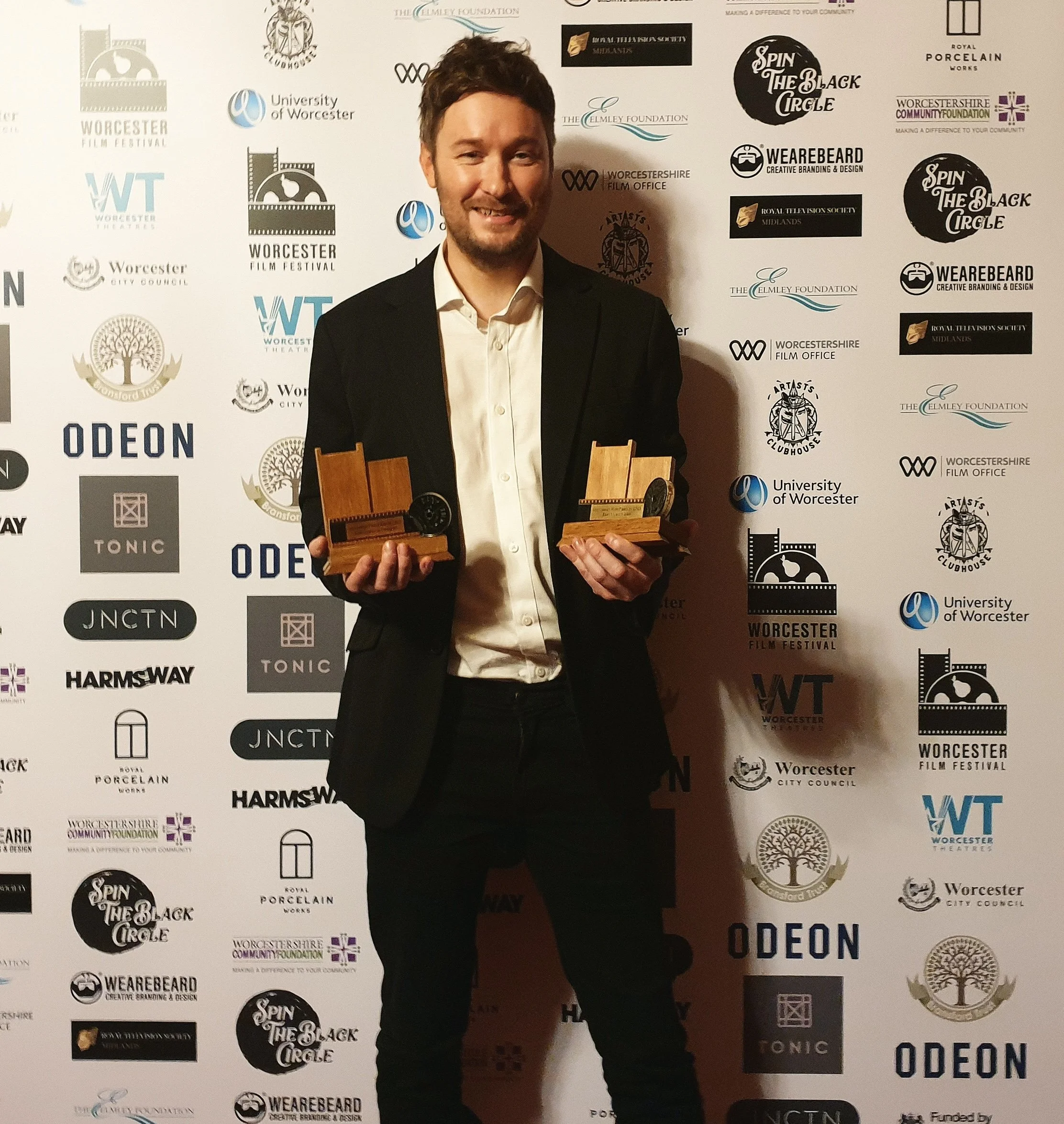 A man in a suit holding two wooden awards, standing in front of a backdrop with multiple logos and event names, smiling at the camera.