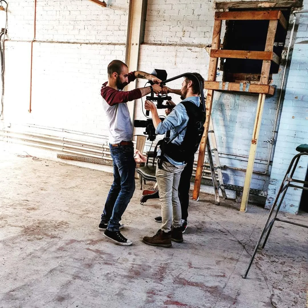 Three men are filming a scene in an industrial setting with exposed brick walls and a wooden structure. One man is holding a boom microphone, another is operating a camera, and the third is assisting.