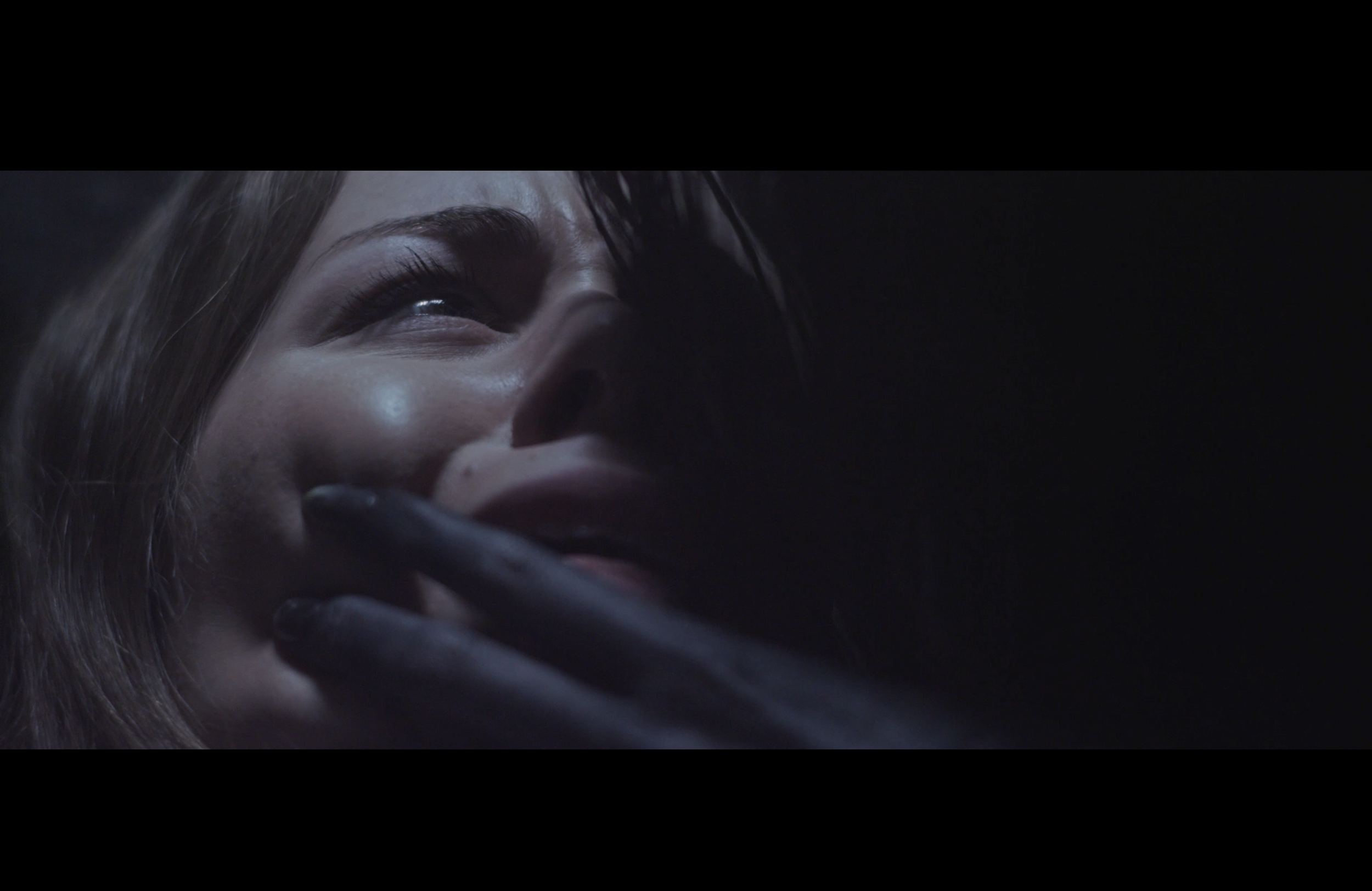 Close-up of a woman with a smile, partially covered by a hand with black gloves, in a dark environment.