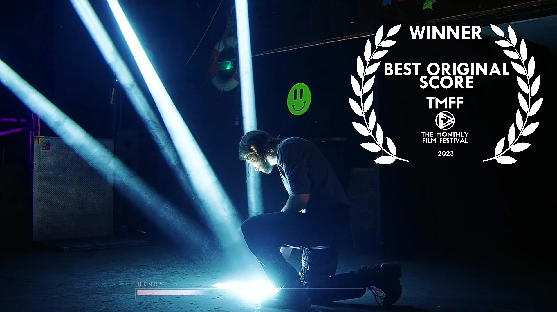 A person crouching on the floor amidst dramatic stage lighting with blue and white beams, dark background, neon green smiley face, and award overlay for best original score at The Monthly Film Festival 2023.