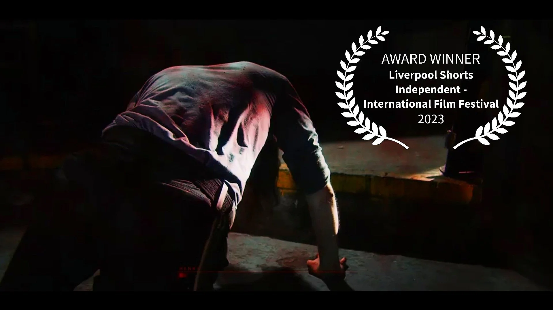 A person kneeling on a surface in a dark environment with a hood over their head, with a glow of light highlighting part of their back and hand. An award laurel wreath displaying 'AWARD WINNER Liverpool Shorts Independent - International Film Festiva