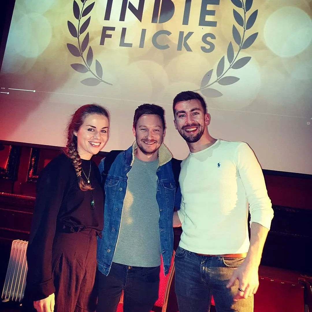 Three people smiling in front of a screen with the words "Indie Flicks" and a laurel wreath design