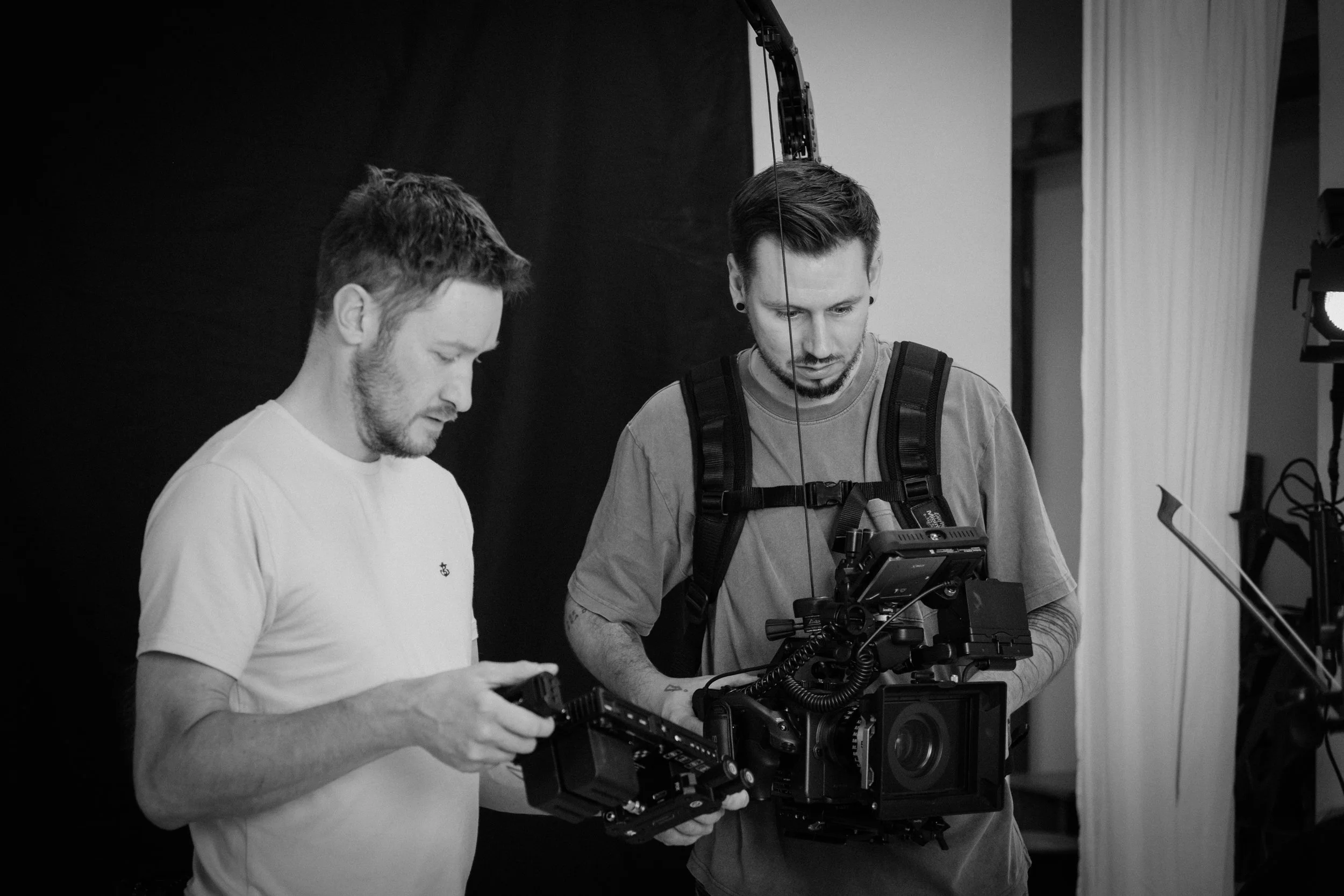 Two men working with video production equipment in a studio, one holding a camera and the other managing a monitor or accessory.