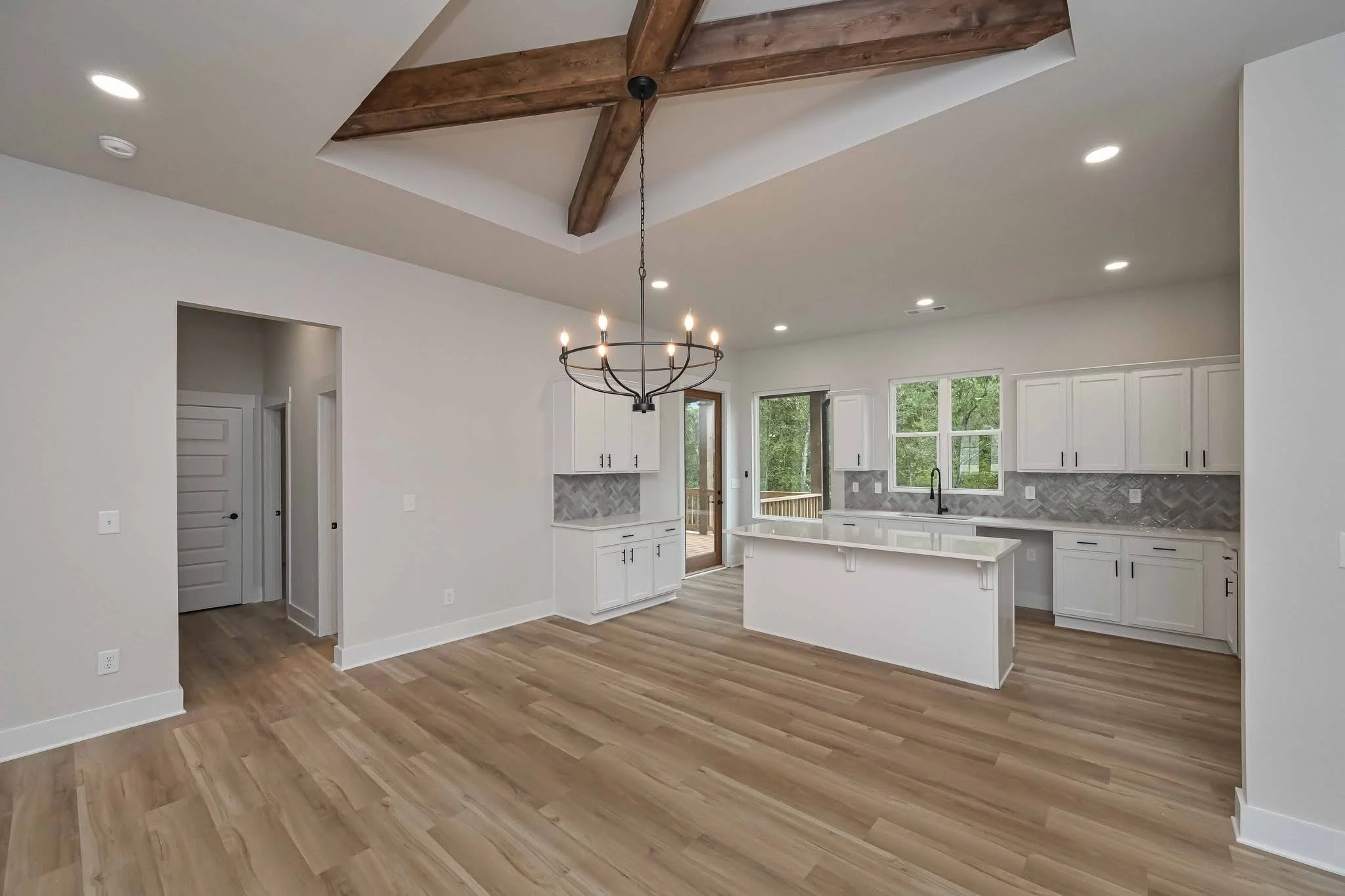 Empty kitchen and dining area with white cabinets, a chandelier, wood flooring, and a ceiling with exposed wooden beams.