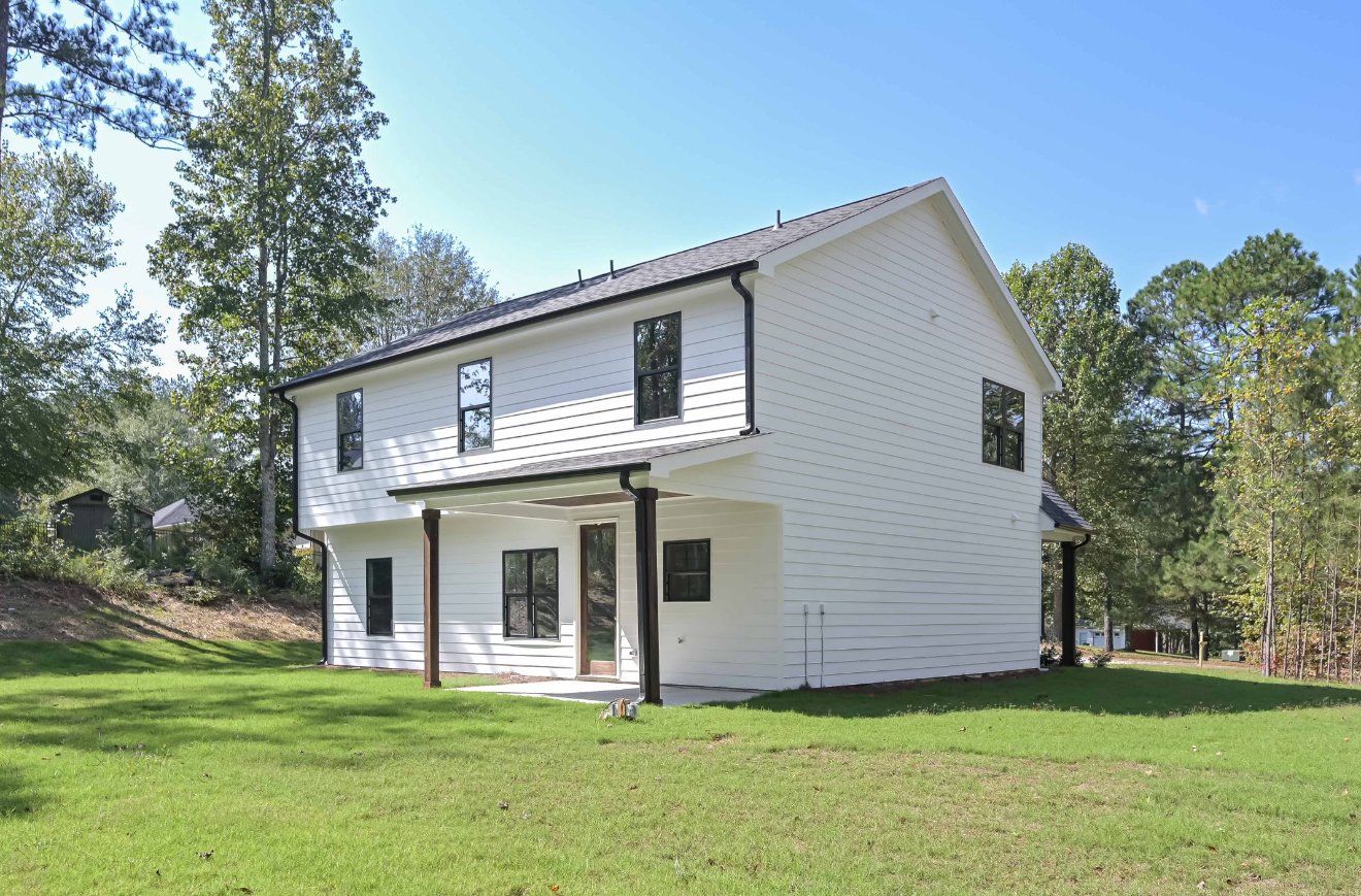 A newly built two-story house with white siding, black window frames, and black gutters, situated on a grassy lawn with trees in the background under a clear blue sky.