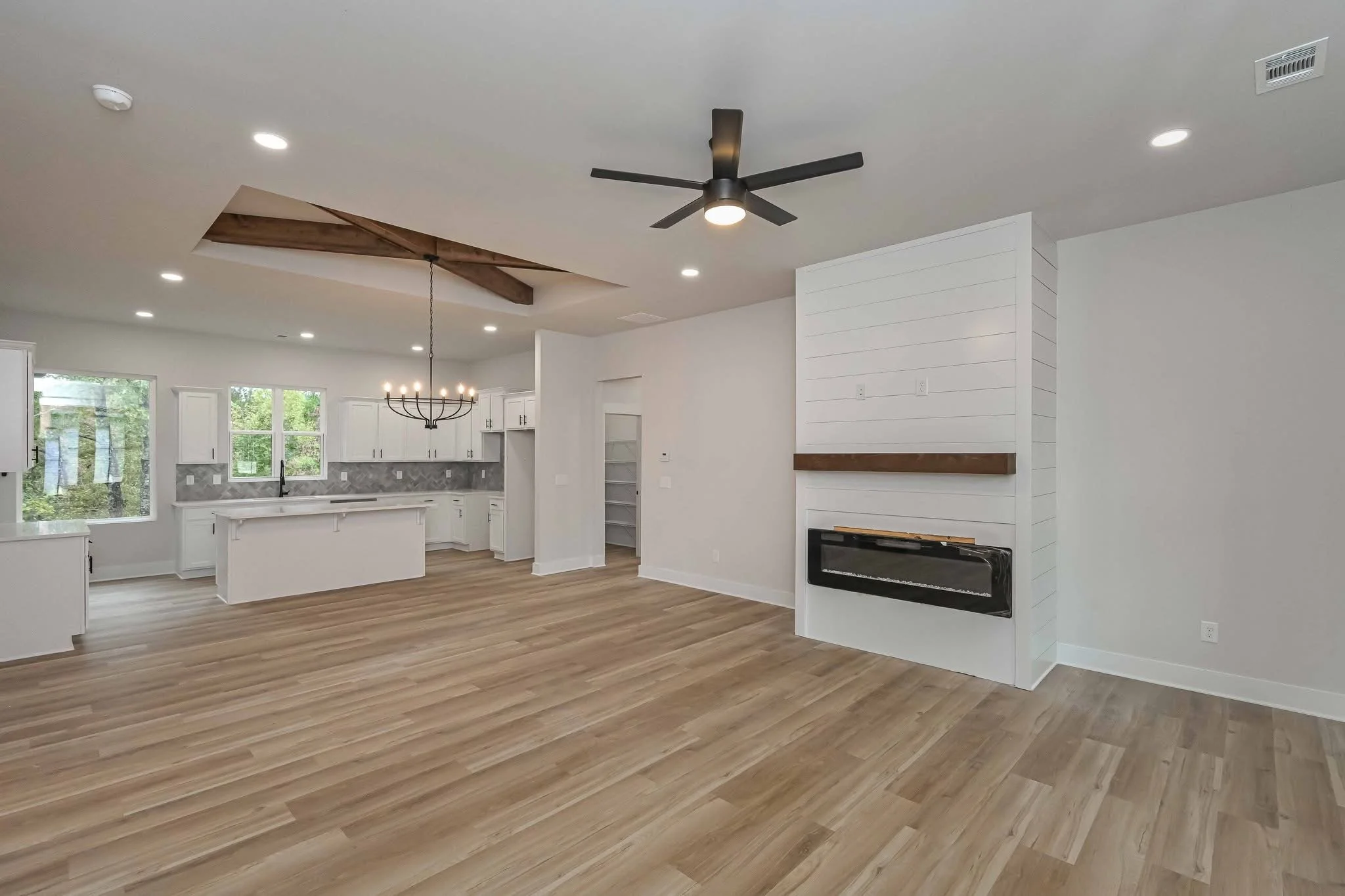 Empty living room with wood flooring, white walls, a black ceiling fan, and a white shiplap fireplace with a wooden mantle. Open kitchen in background with white cabinetry, gray tile backsplash, island, and a chandelier.