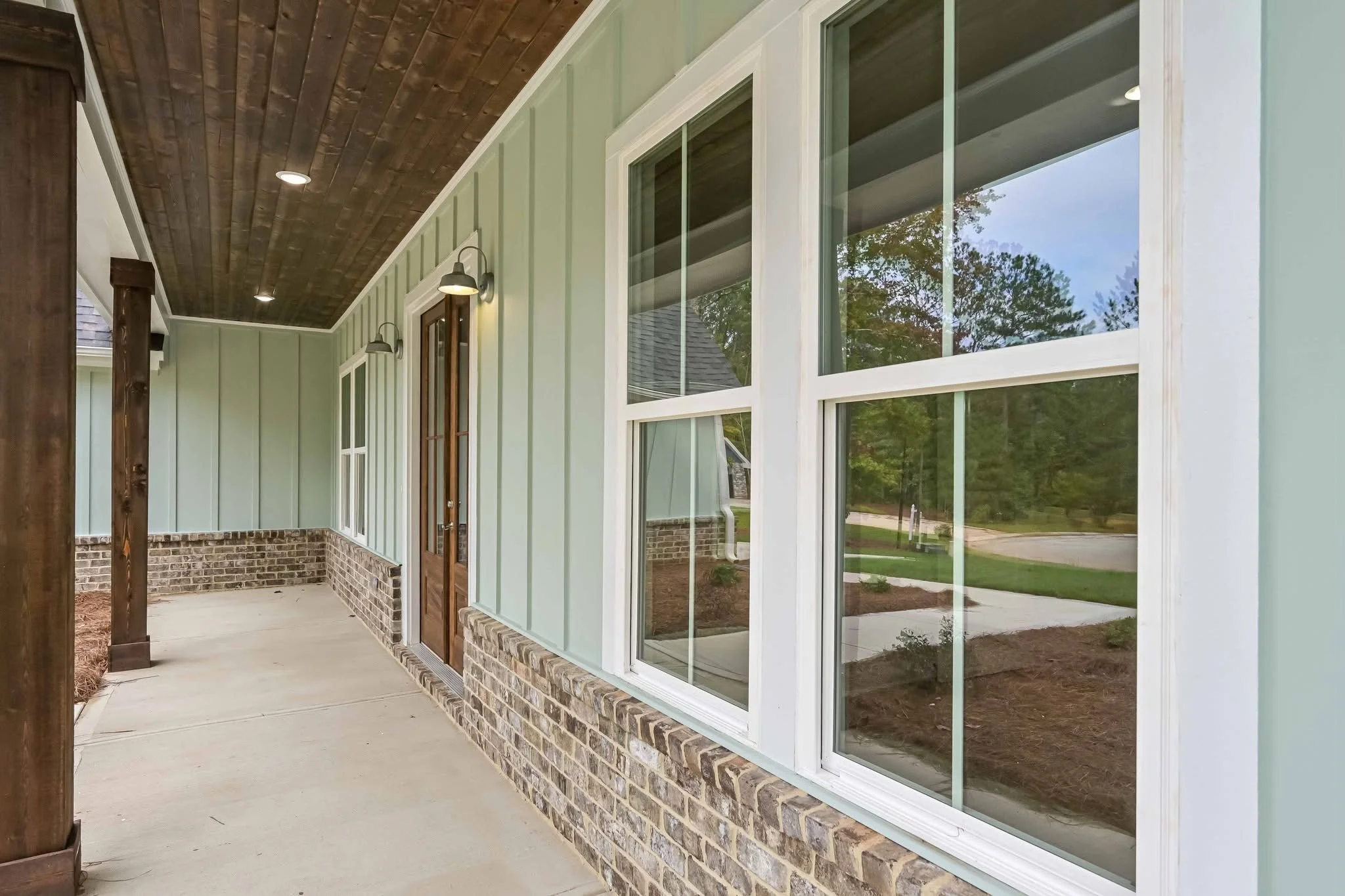 View of a house porch with green siding, brick base, wooden front door, and large double-hung window reflecting trees and a sidewalk outside.