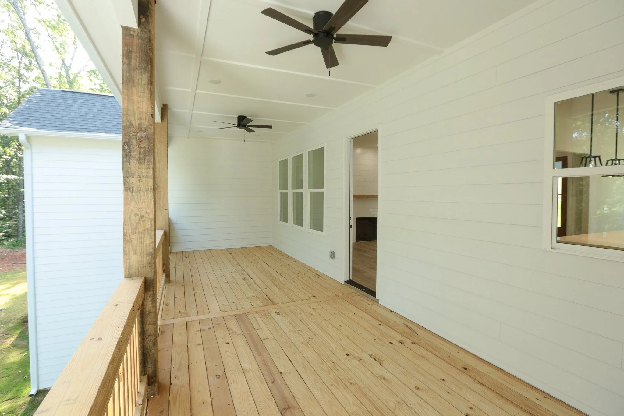 Empty covered porch with wooden floor, white walls, and two ceiling fans, attached to a house with a sliding door and windows.