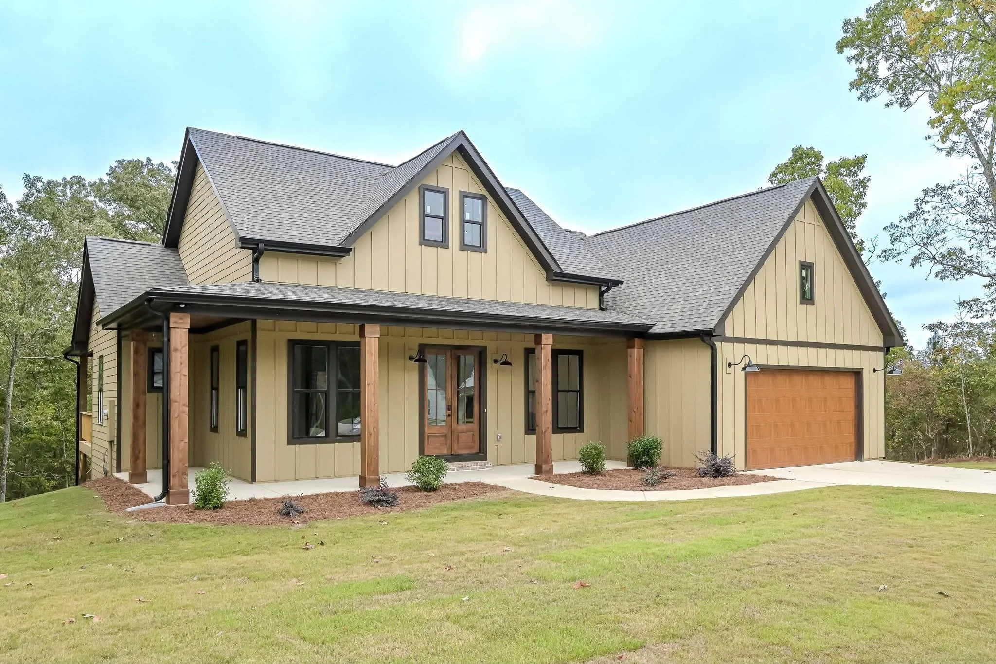 A modern two-story house with beige vertical siding, black window frames, a wooden front door, a two-car garage with wood-textured door, and a front lawn with small shrubs, set against a clear sky and trees in the background.